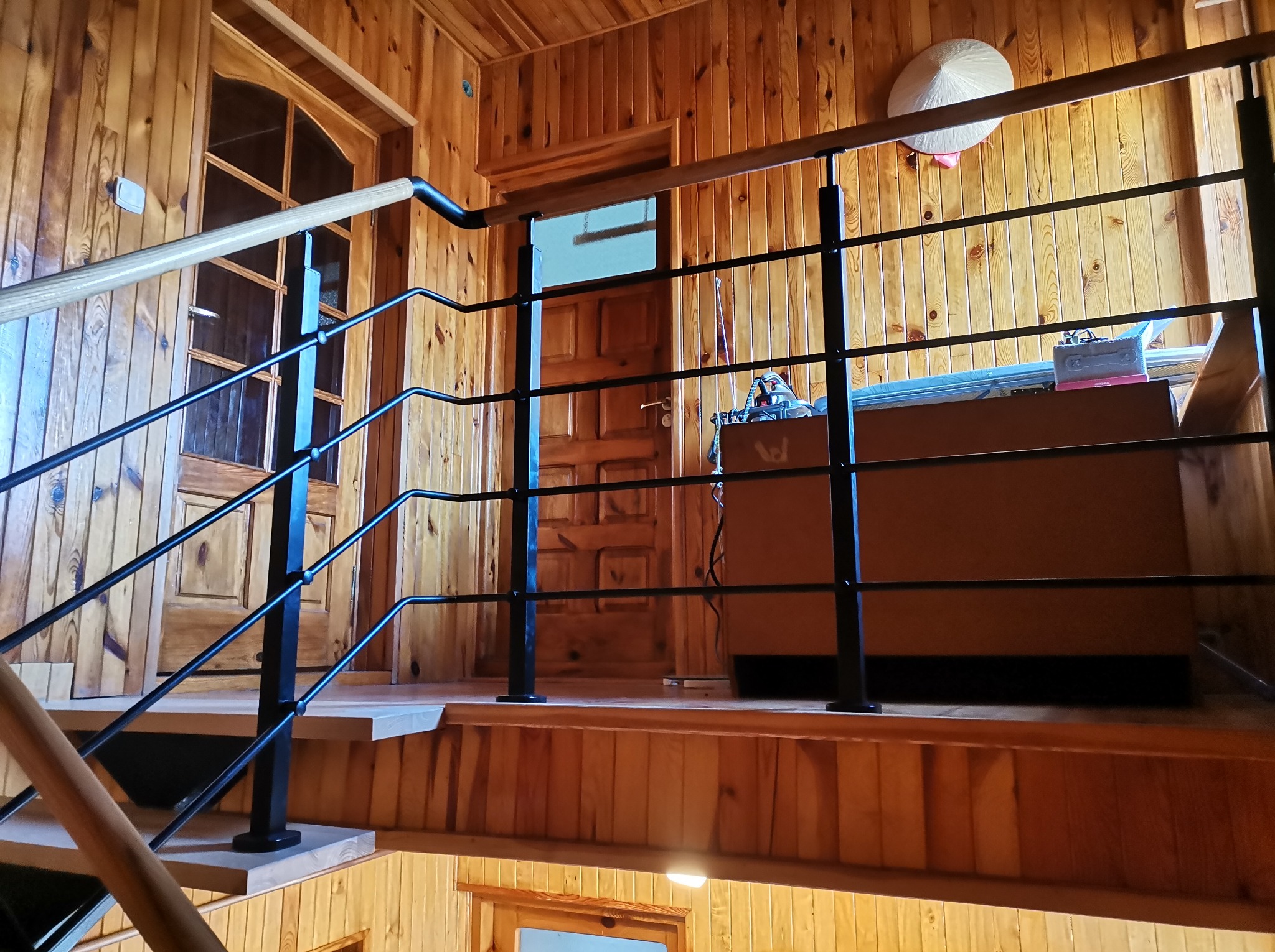 Wnętrze domu z drewnianą boazerią i czarną, metalową balustradą schodową. Na ścianie wisi słomkowy kapelusz. Widoczne drewniane schody i drzwi.