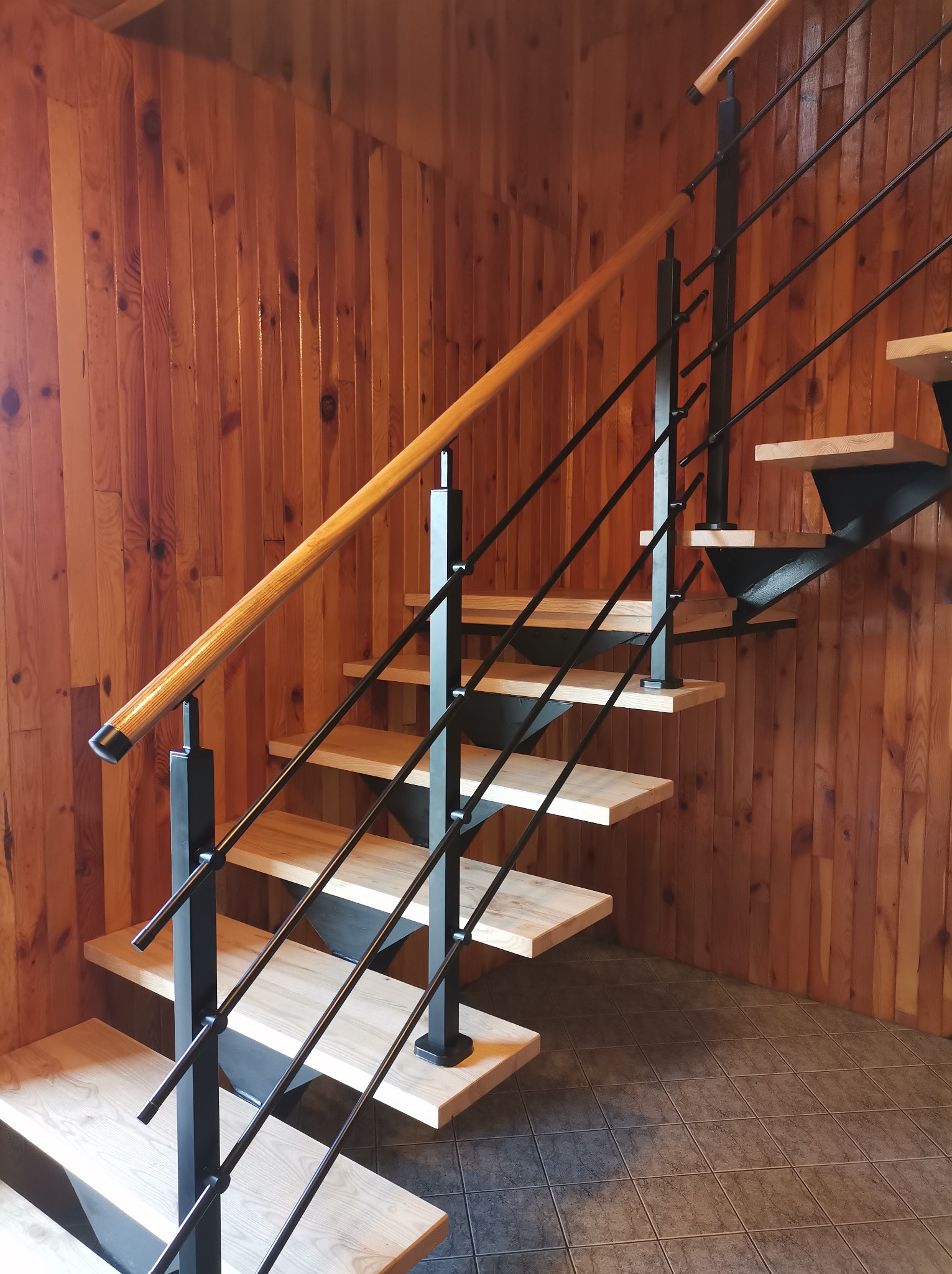 Nowoczesne schody z drewnianymi stopniami i czarną metalową balustradą na tle drewnianej ściany. Jasne drewno i ciemny metal tworzą kontrast.