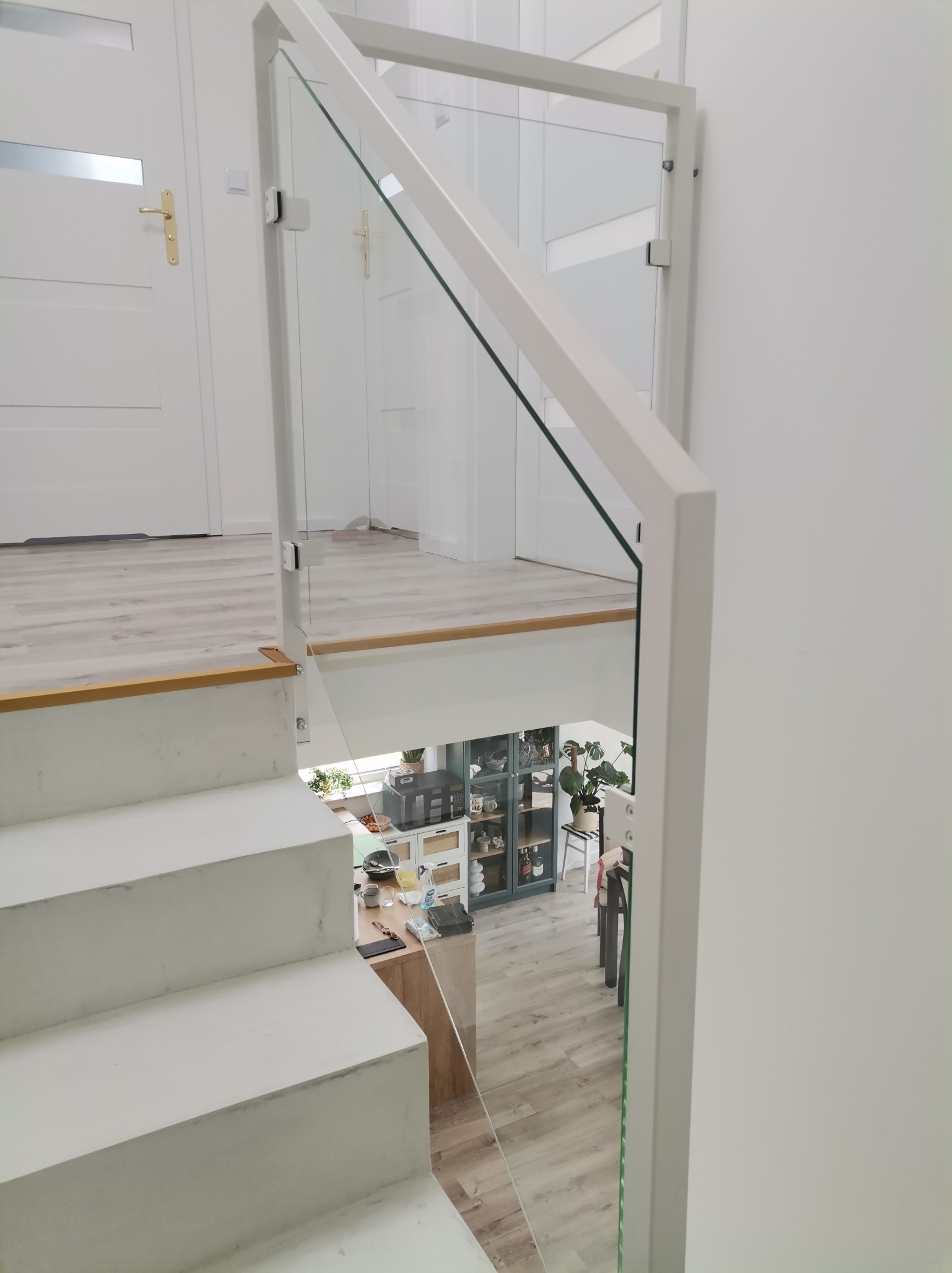 Nowoczesna, minimalistyczna balustrada szklana z białymi elementami konstrukcyjnymi, zamontowana przy schodach w jasnym wnętrzu. Widok z góry na dół.