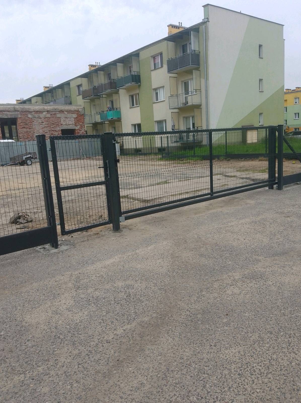 Nowoczesna, przesuwna brama panelowa w kolorze grafitowym, z widocznym mechanizmem otwierania, na tle bloku mieszkalnego i fragmentu starej zabudowy.