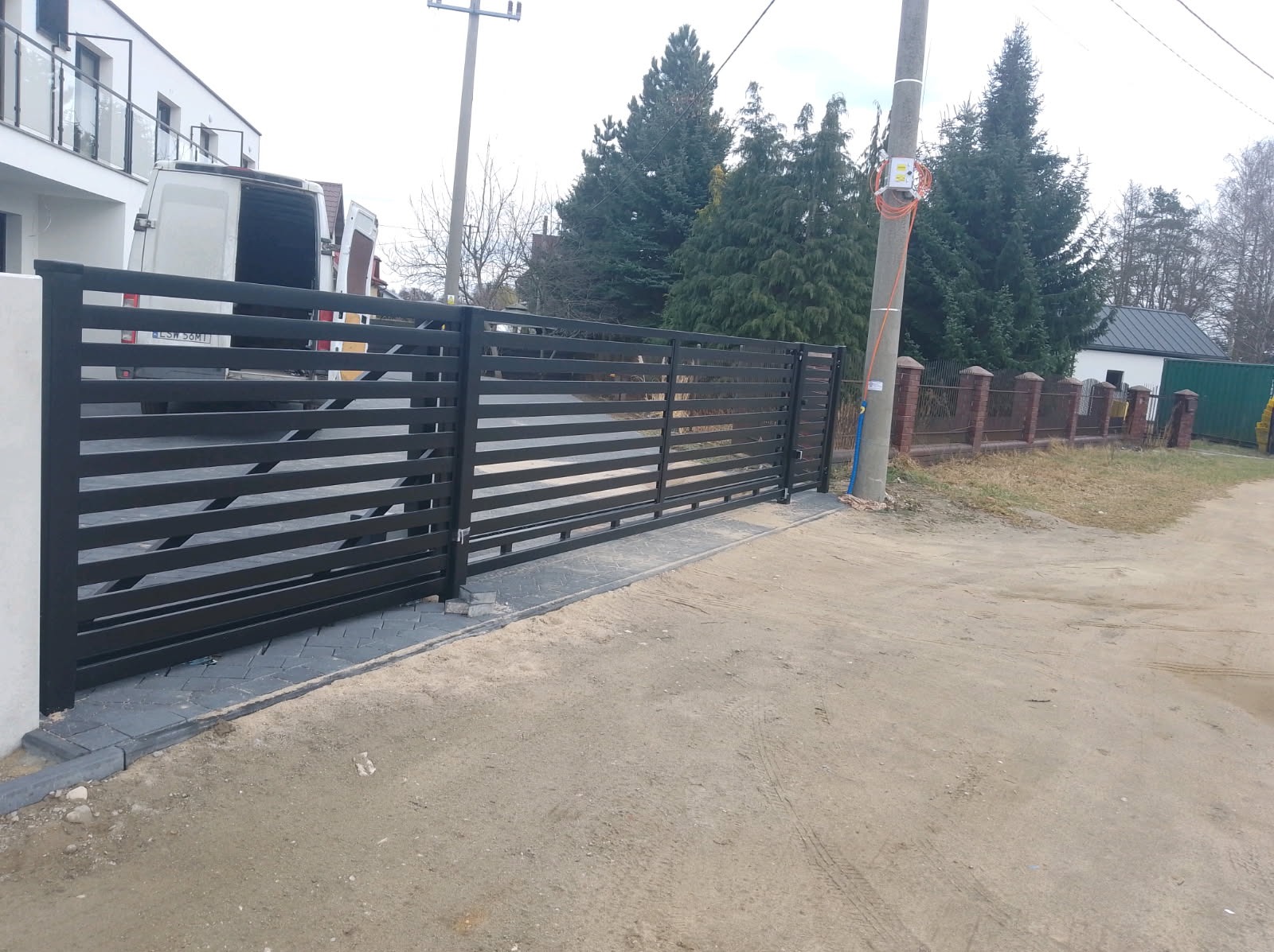 Nowoczesna, pozioma brama przesuwna w kolorze antracytowym, z widocznym fragmentem posesji i zaparkowanym busem w tle. Brama zamontowana na kostce brukowej.