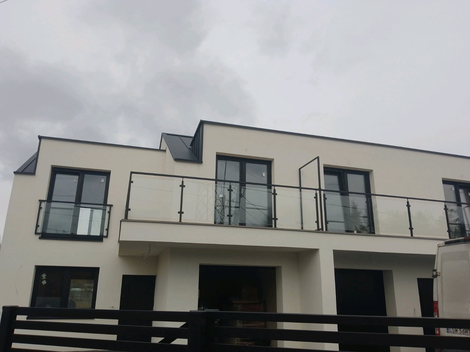 Nowoczesny dom z balkonami zabezpieczonymi szklanymi balustradami z czarnymi elementami. W tle pochmurne niebo i fragment ogrodzenia panelowego.