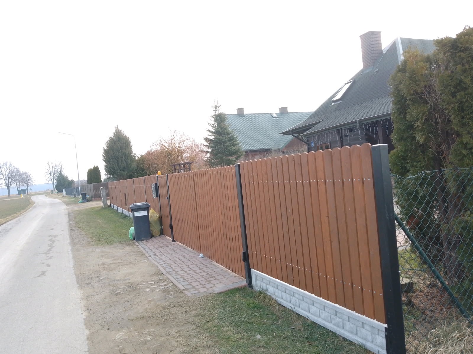 Nowoczesne ogrodzenie panelowe z brązowych desek z podmurówką z szarej cegły, widok z boku na tle wiejskiego krajobrazu i domu z zielonym dachem.