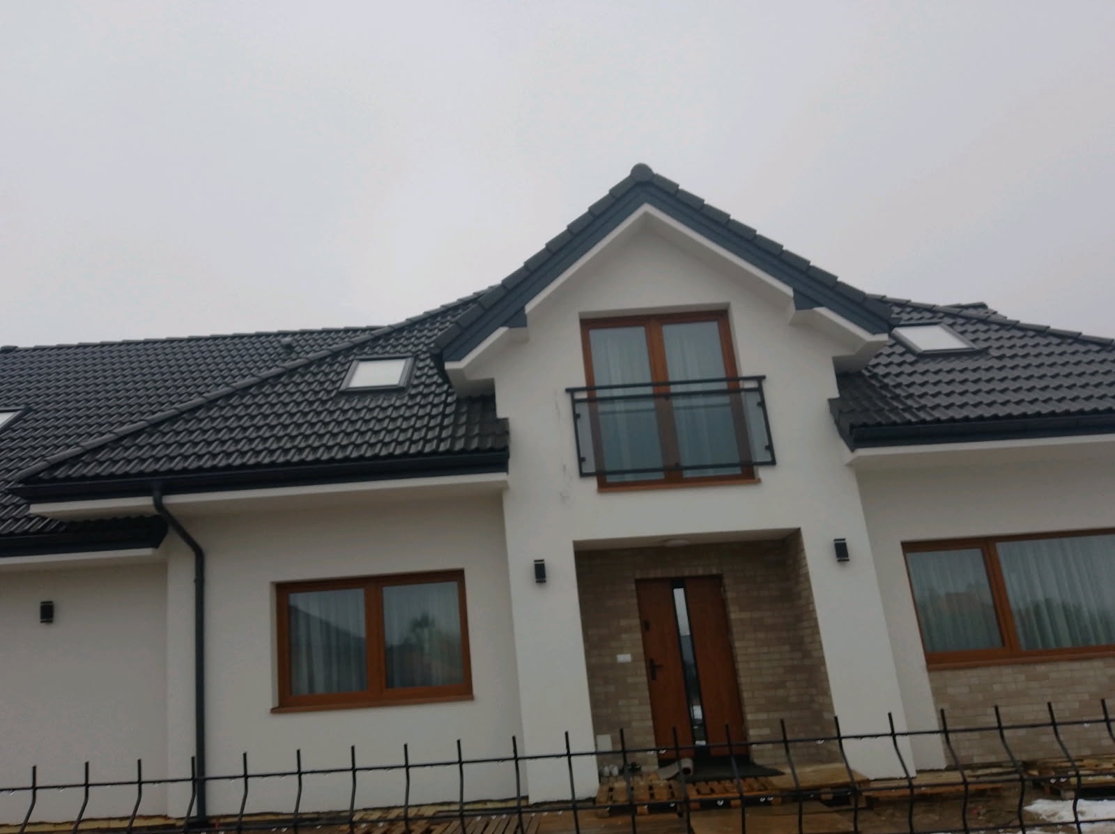 Nowoczesny dom z czarną dachówką i metalowym ogrodzeniem panelowym. Widoczne okna z drewnianą ramą i balkon z szklaną balustradą. Pochmurne niebo w tle.