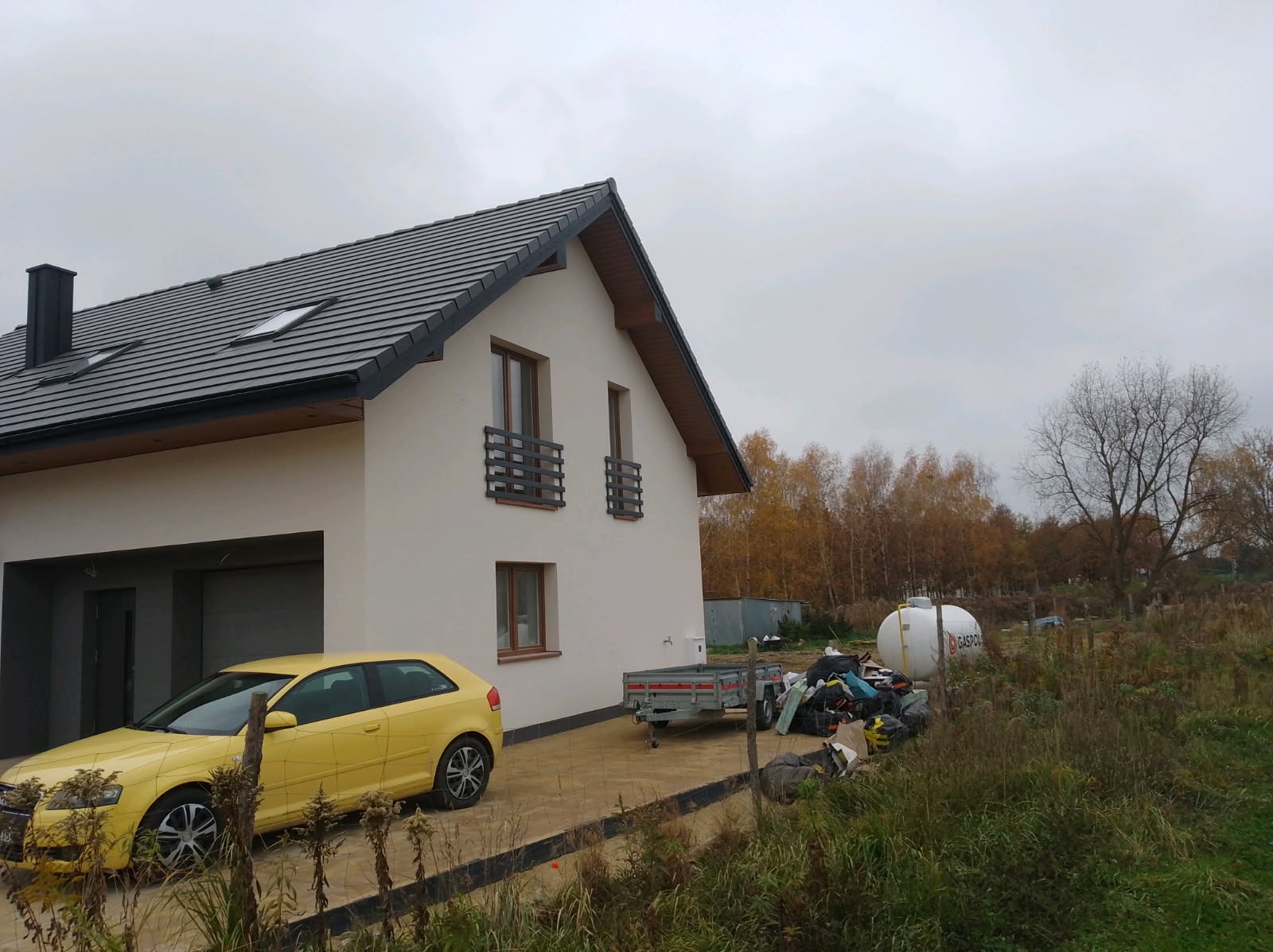 Nowoczesny dom z szarą dachówką i żółtym autem. Widoczne ogrodzenie posesji, zbiornik na gaz i przyczepka. Jesienna sceneria z drzewami w tle.