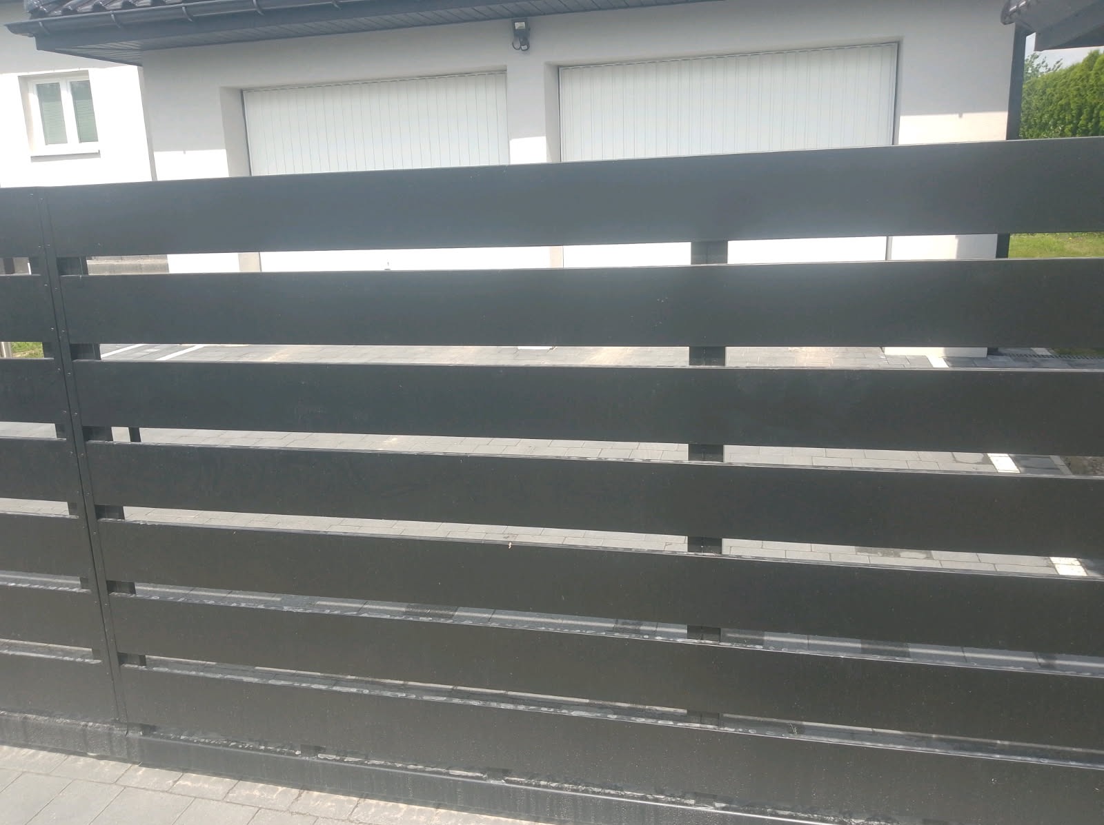 Nowoczesne, poziome przęsła ogrodzeniowe w kolorze grafitowym, na tle białego budynku z dwoma bramami garażowymi. Minimalistyczny design, widok z bliska.