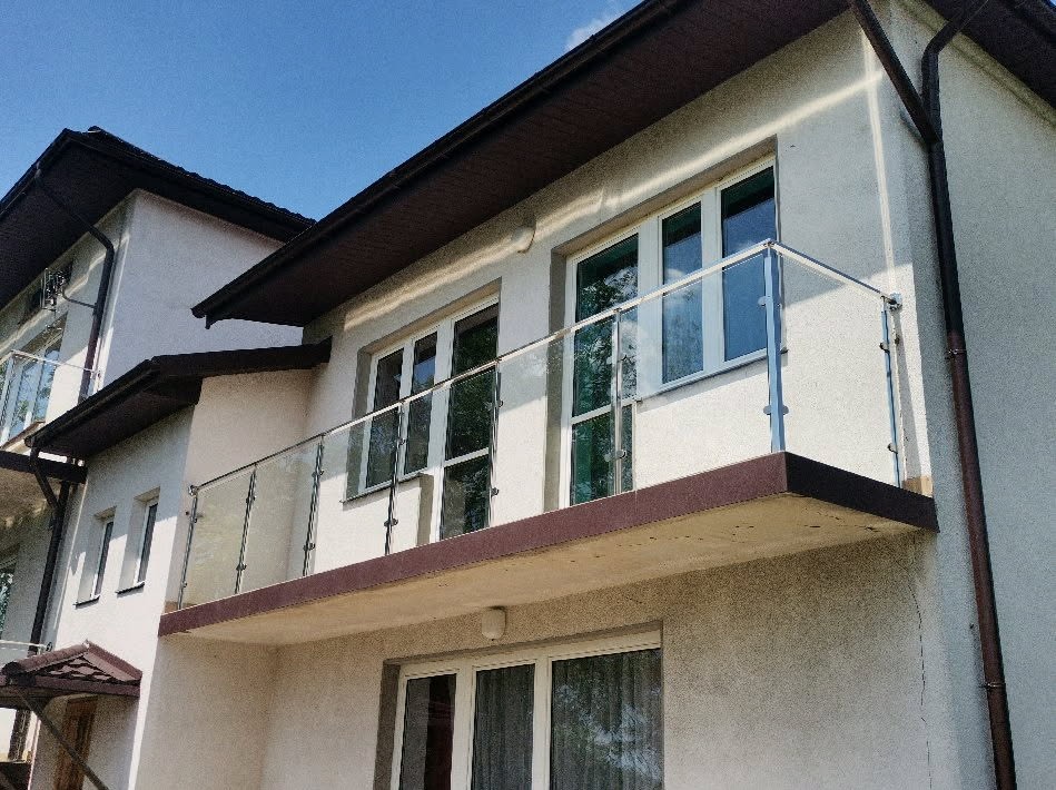 Nowoczesna balustrada balkonowa ze szkła i stali nierdzewnej, zamontowana na elewacji dwupiętrowego domu z brązowym dachem. Widok z dołu, błękitne niebo w tle.