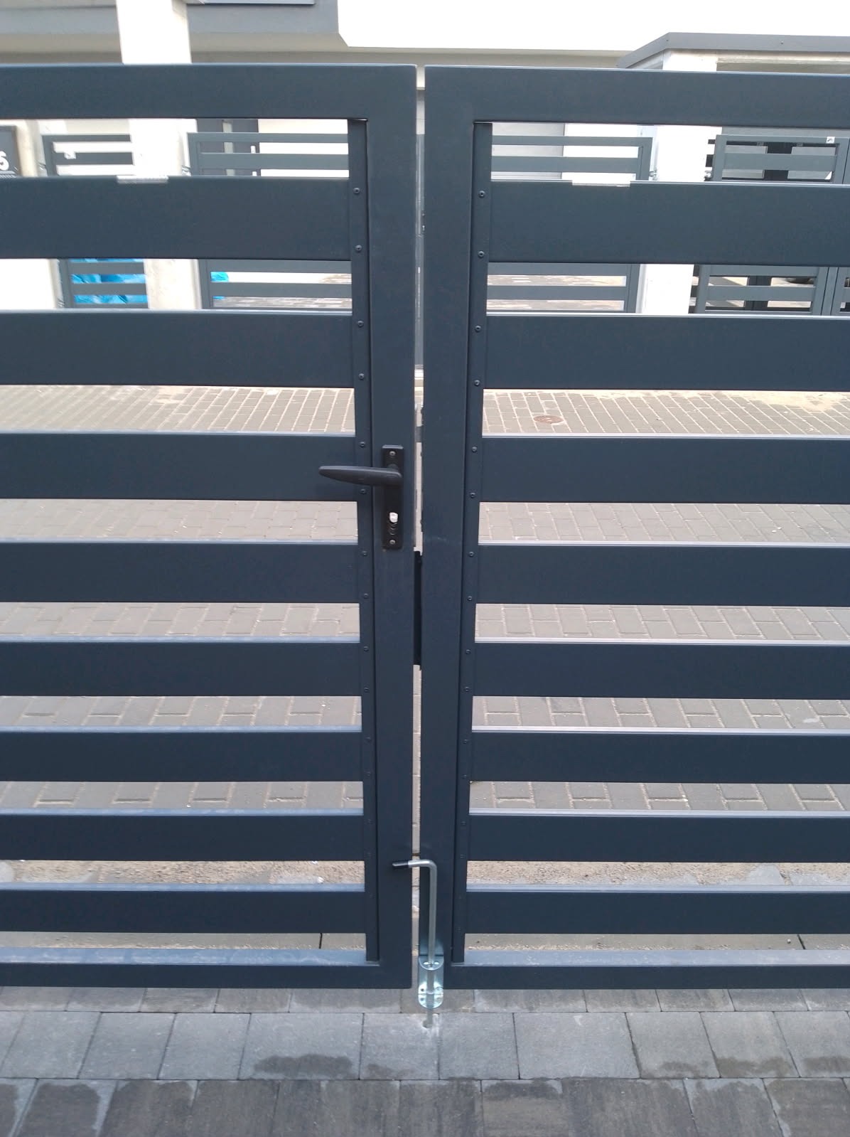 Nowoczesna, grafitowa brama posesyjna z poziomymi panelami, czarną klamką i metalowym ryglem, zamontowana na kostce brukowej. Minimalistyczny design.