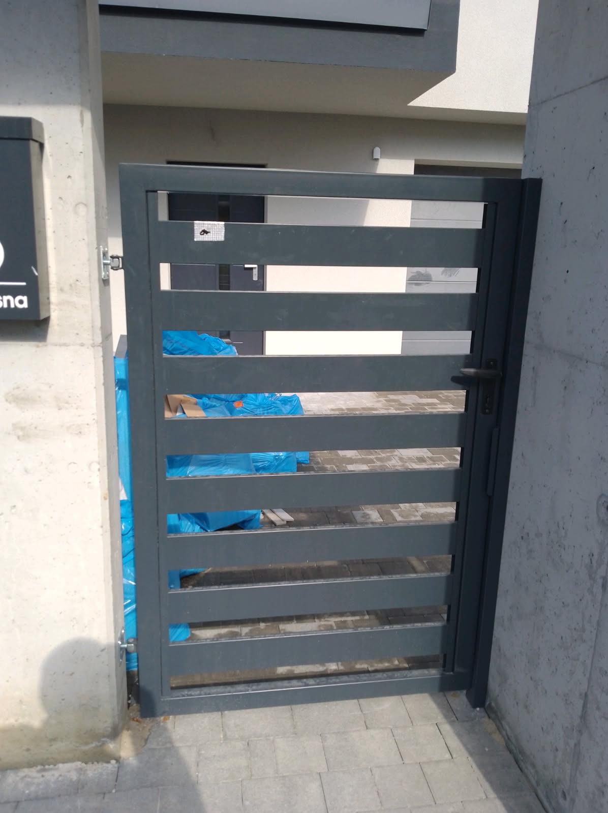 Nowoczesna, grafitowa furtka z poziomych profili, zamontowana przy betonowym słupku. W tle widoczne materiały budowlane zabezpieczone folią.