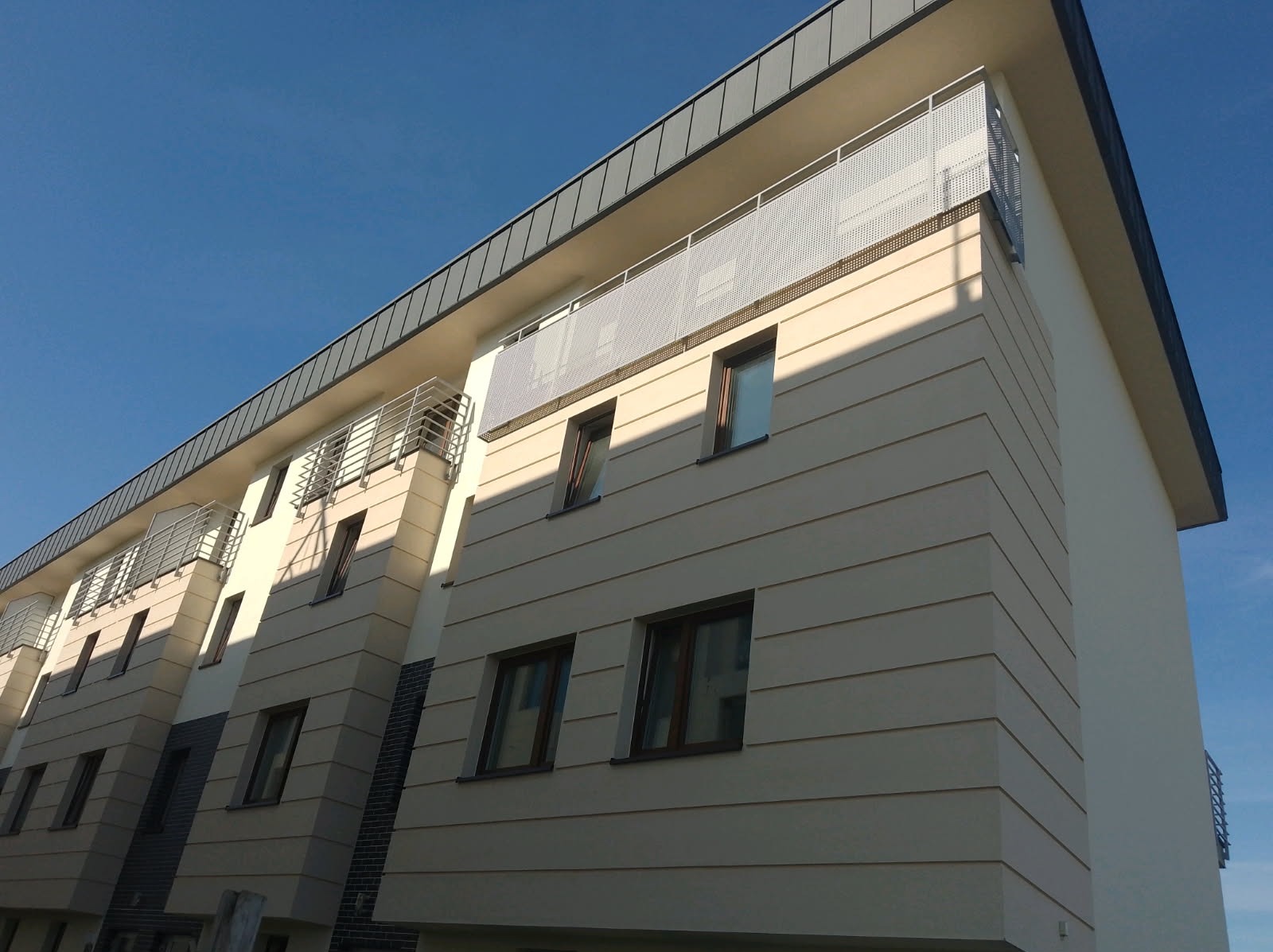 Nowoczesny budynek wielorodzinny z balkonami zabezpieczonymi metalowymi, ażurowymi balustradami. Jasna elewacja z poziomymi pasami i ciemne okna.