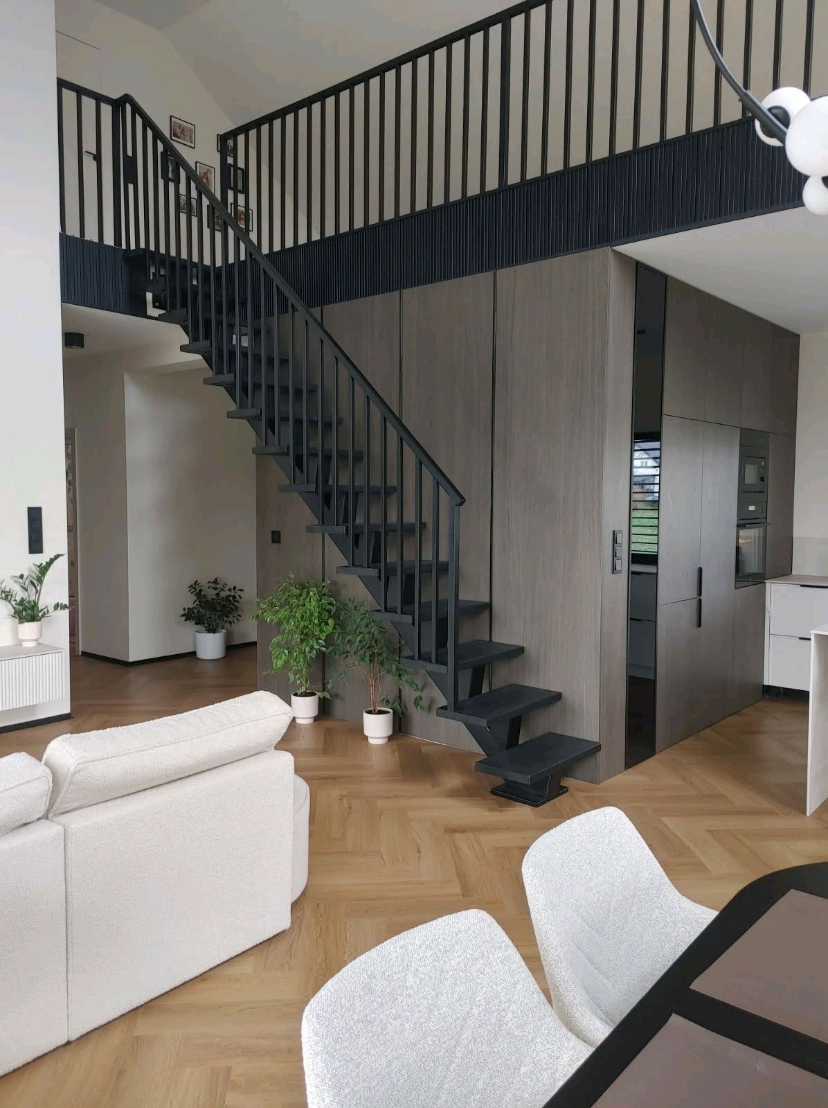 Nowoczesne, czarne schody z metalową balustradą w jasnym wnętrzu domu. Minimalistyczny design, drewniana podłoga, rośliny doniczkowe. Ujęcie z dołu, widok na schody i antresolę.