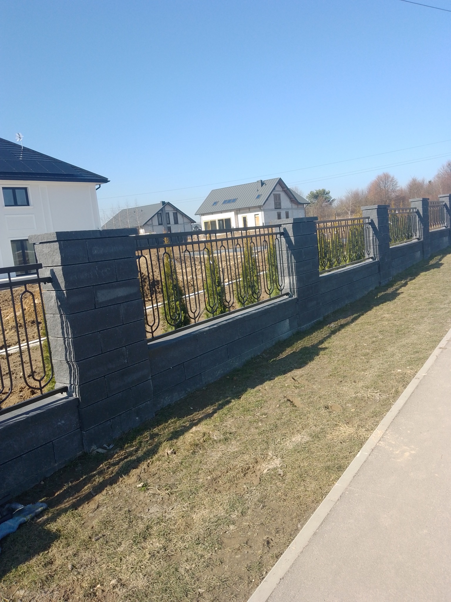 Nowoczesne ogrodzenie z szarej kostki i metaloplastyki, oddzielające posesję z tujami od chodnika. W tle domy jednorodzinne w słoneczny dzień.