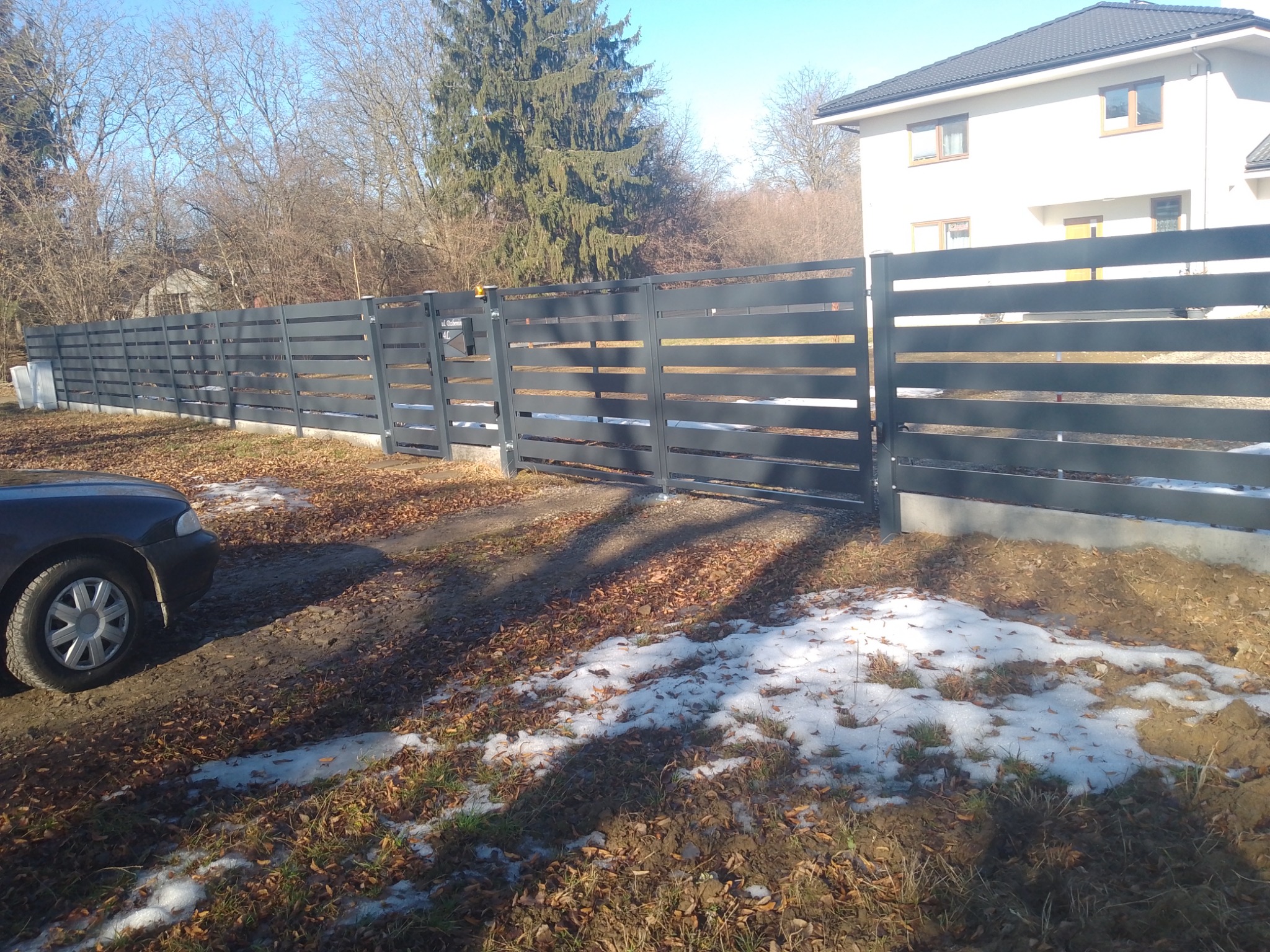 Nowoczesne, poziome ogrodzenie panelowe w kolorze grafitowym, z bramą wjazdową, na tle domu i drzew bez liści, z resztkami śniegu na ziemi.