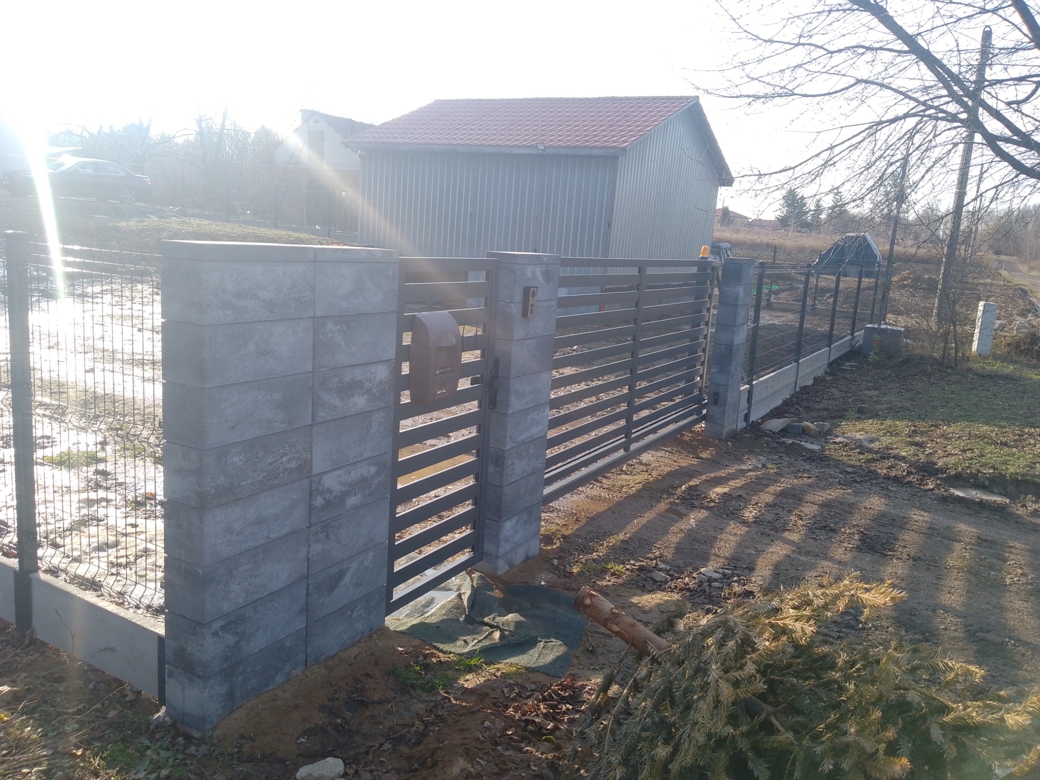 Nowoczesne ogrodzenie panelowe z betonowymi słupkami i furtką, skrzynka na listy. W tle budynek gospodarczy i drzewa bez liści, słoneczny dzień.