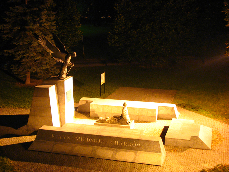 Nocne ujęcie pomnika ofiar Katynia, Miednoje i Charkowa z figurą anioła na szczycie i klęczącą postacią na dole, oświetlonego reflektorami.