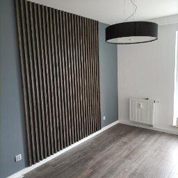 PM wykonczenie wnetrz - Nowoczesne wykończenie wnętrza: drewniane lamele na ścianie w odcieniach brązu, szara ściana, podłoga imitująca drewno i okrągła lampa sufitowa.