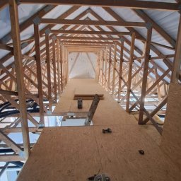 PM wykonczenie wnetrz - Szeroki kadr na drewnianą konstrukcję dachu z płytami OSB, widok z dołu. Na płytach leżą narzędzia i akcesoria montażowe. W tle widoczna drabina i oświetlenie.