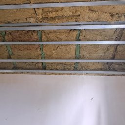 PM wykonczenie wnetrz - Montaż izolacji z wełny mineralnej na metalowym ruszcie sufitowym, widoczne zielone elementy mocujące. Ściana w tle przygotowana do dalszych prac wykończeniowych.