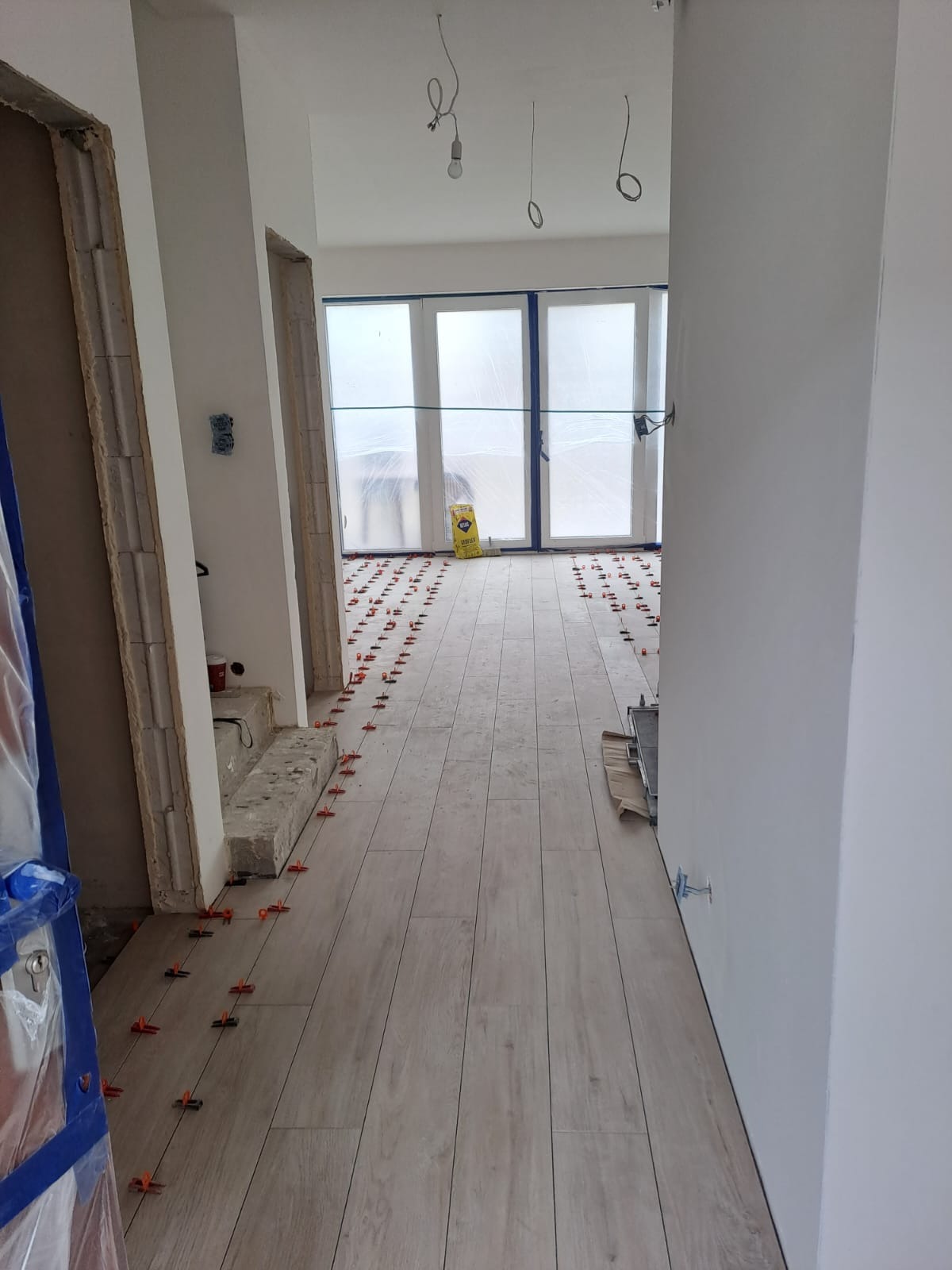 Jasne płytki podłogowe ułożone w korytarzu, z użyciem klipsów poziomujących. Widoczne przygotowania do dalszych prac wykończeniowych, okna zasłonięte folią.