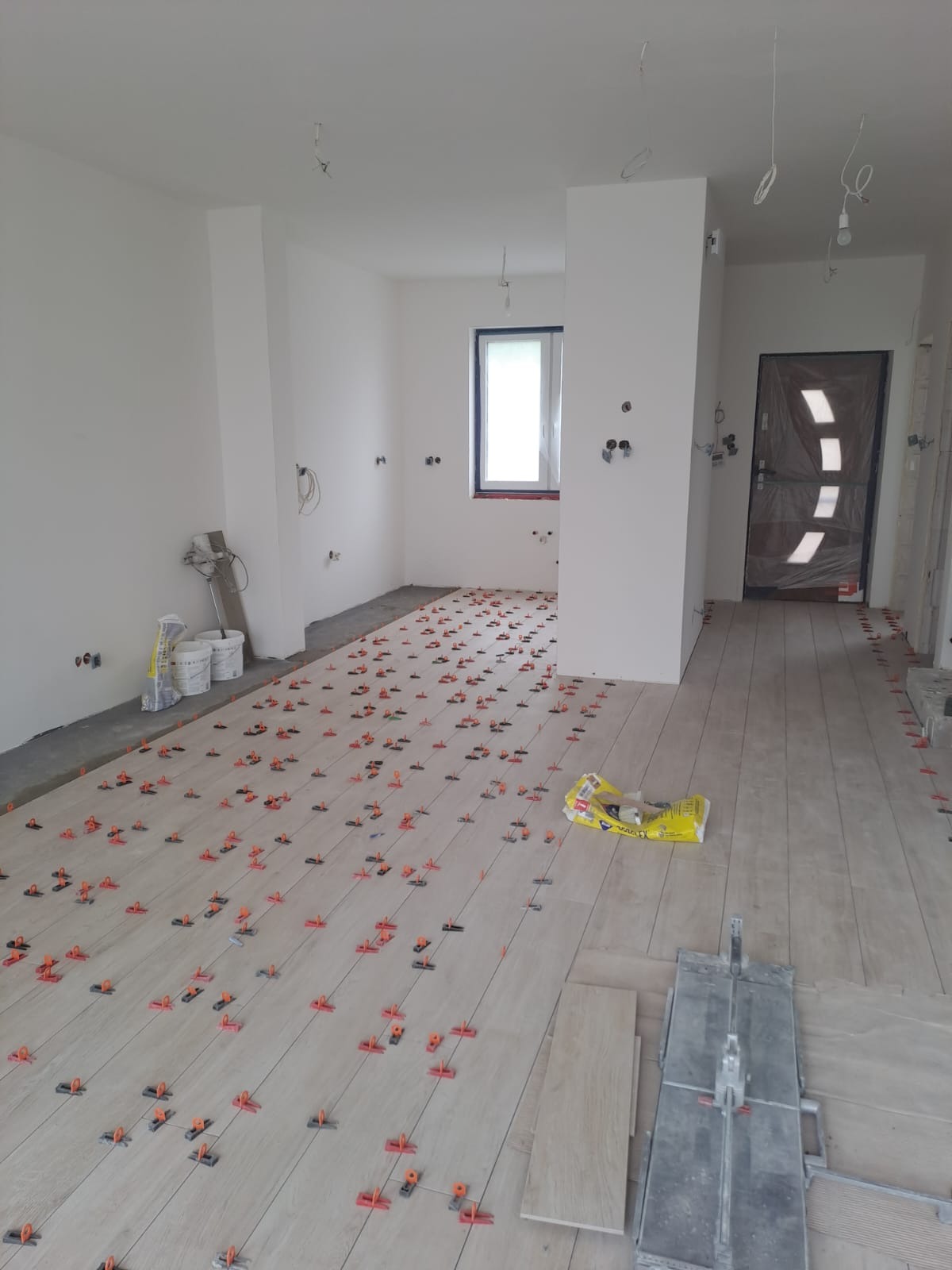 Układanie płytek podłogowych w jasnym odcieniu drewna, z użyciem klipsów poziomujących. Widoczna przecinarka do płytek i worki z zaprawą. W tle surowe ściany i okno.