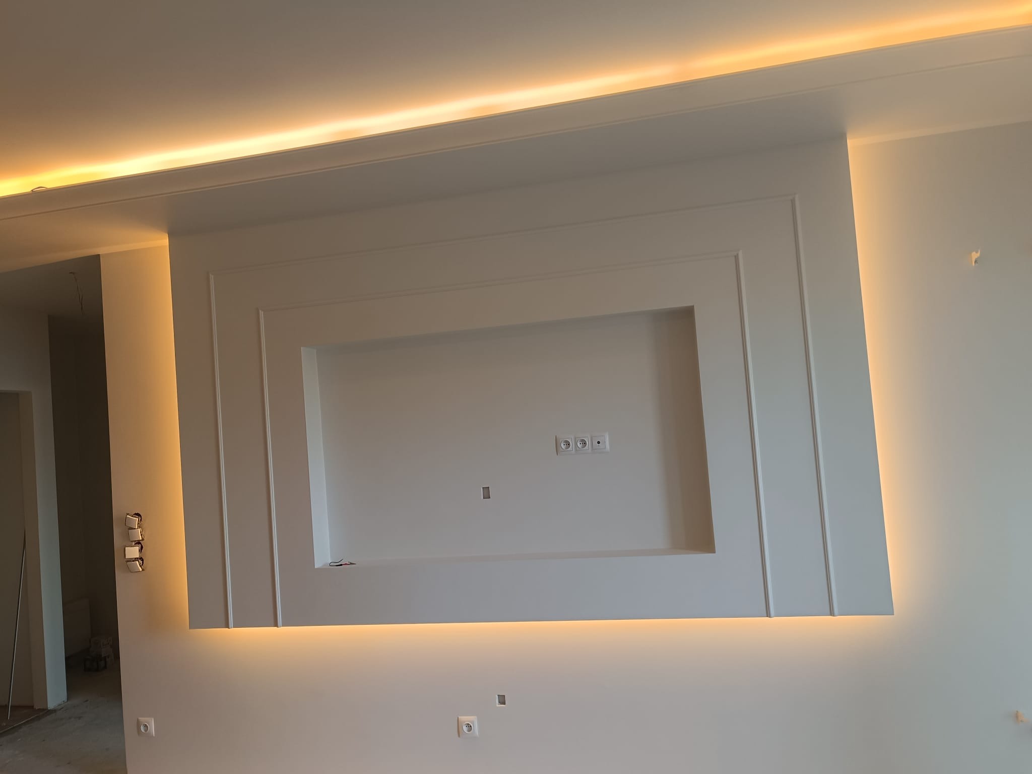 Biała ściana z dekoracyjną wnęką podświetloną ciepłym światłem LED. Widoczne gniazdka elektryczne i włączniki. Minimalistyczny design wnętrza.