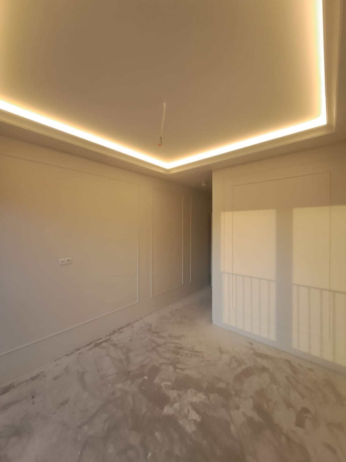 Wykończone wnętrze pokoju z dekoracyjnymi listwami ściennymi i podświetlanym sufitem. Podłoga przygotowana do dalszych prac wykończeniowych.