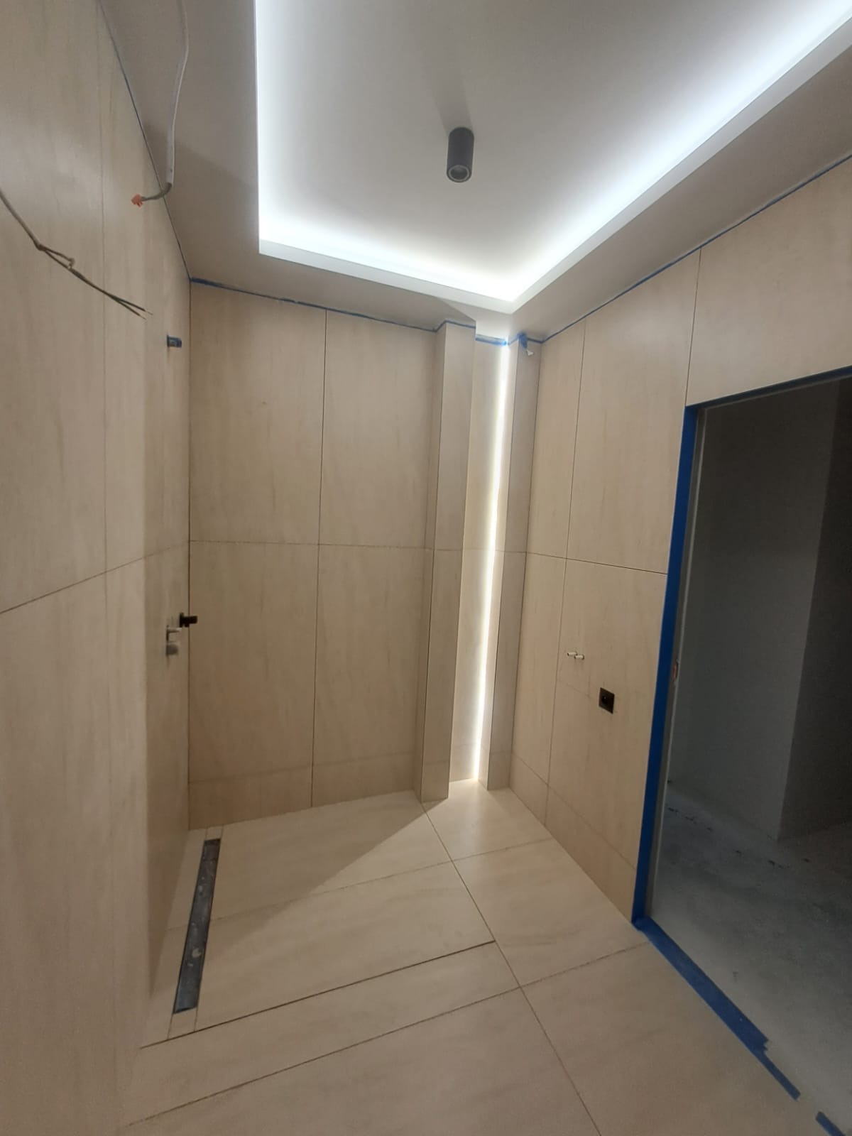 Wykończona łazienka z beżowymi płytkami, oświetleniem LED na suficie i pionowym paskiem światła. Widoczny odpływ liniowy i zabezpieczone taśmą malarską obramowanie drzwi.