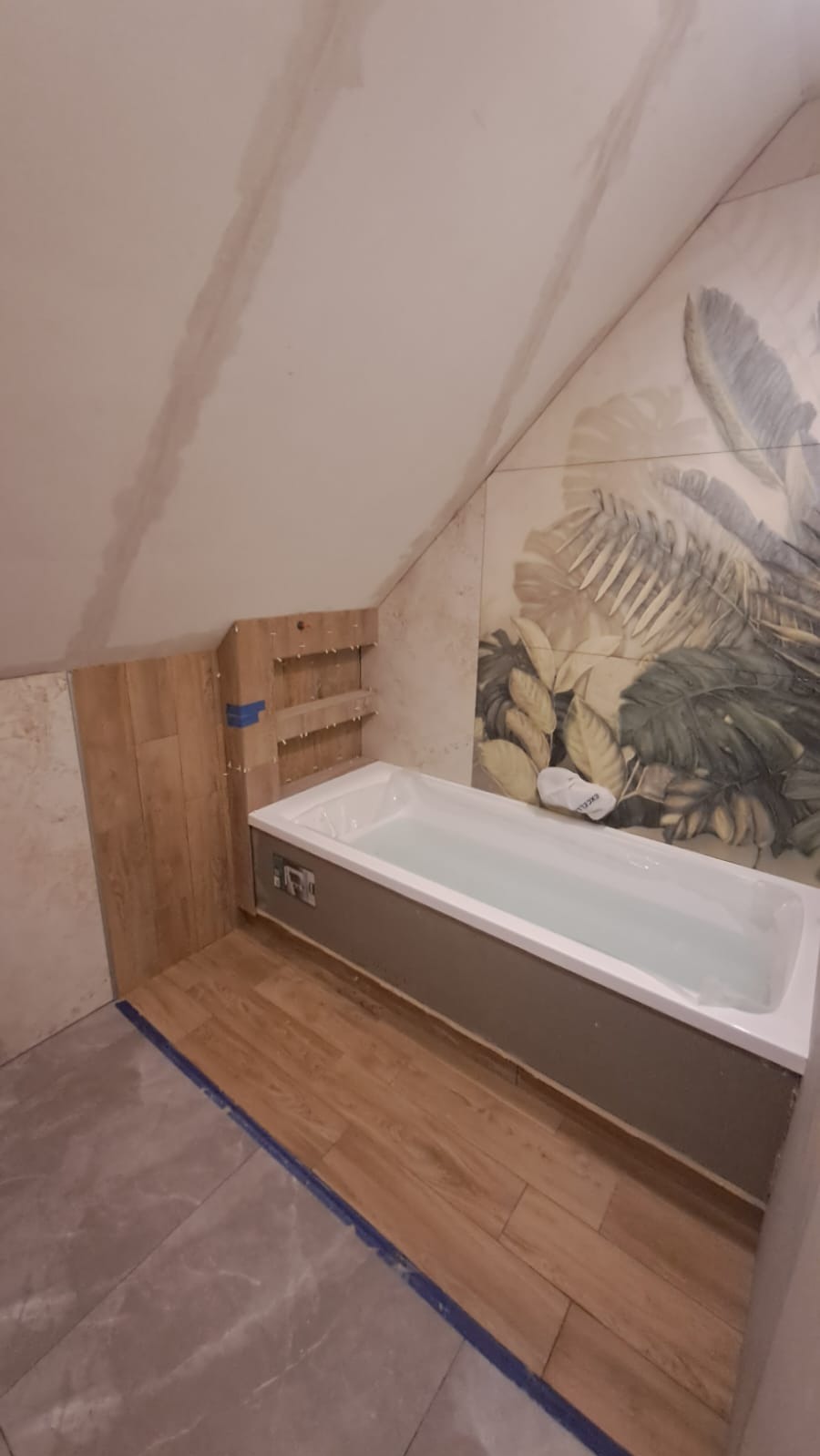 Łazienka na poddaszu w trakcie wykończenia: wanna wypełniona wodą, częściowo ułożone płytki imitujące drewno, motyw roślinny na ścianie. Widoczne krzyżyki glazurnicze.
