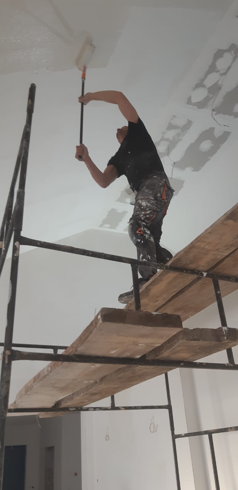 Pracownik na rusztowaniu maluje sufit wałkiem w Zielonej Górze. Widoczne ślady po szpachlowaniu i niedomalowane fragmenty. Ubranie robocze z plamami farby.