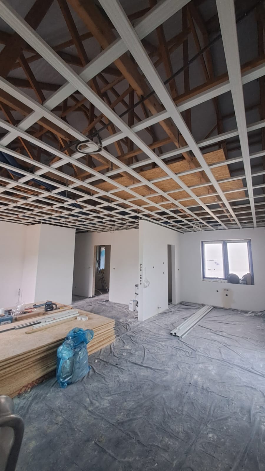 Sufit podwieszany z metalową konstrukcją w trakcie montażu. Widoczne płyty OSB, narzędzia i materiały budowlane na podłodze zabezpieczonej folią. Stan surowy, okno z parapetem.