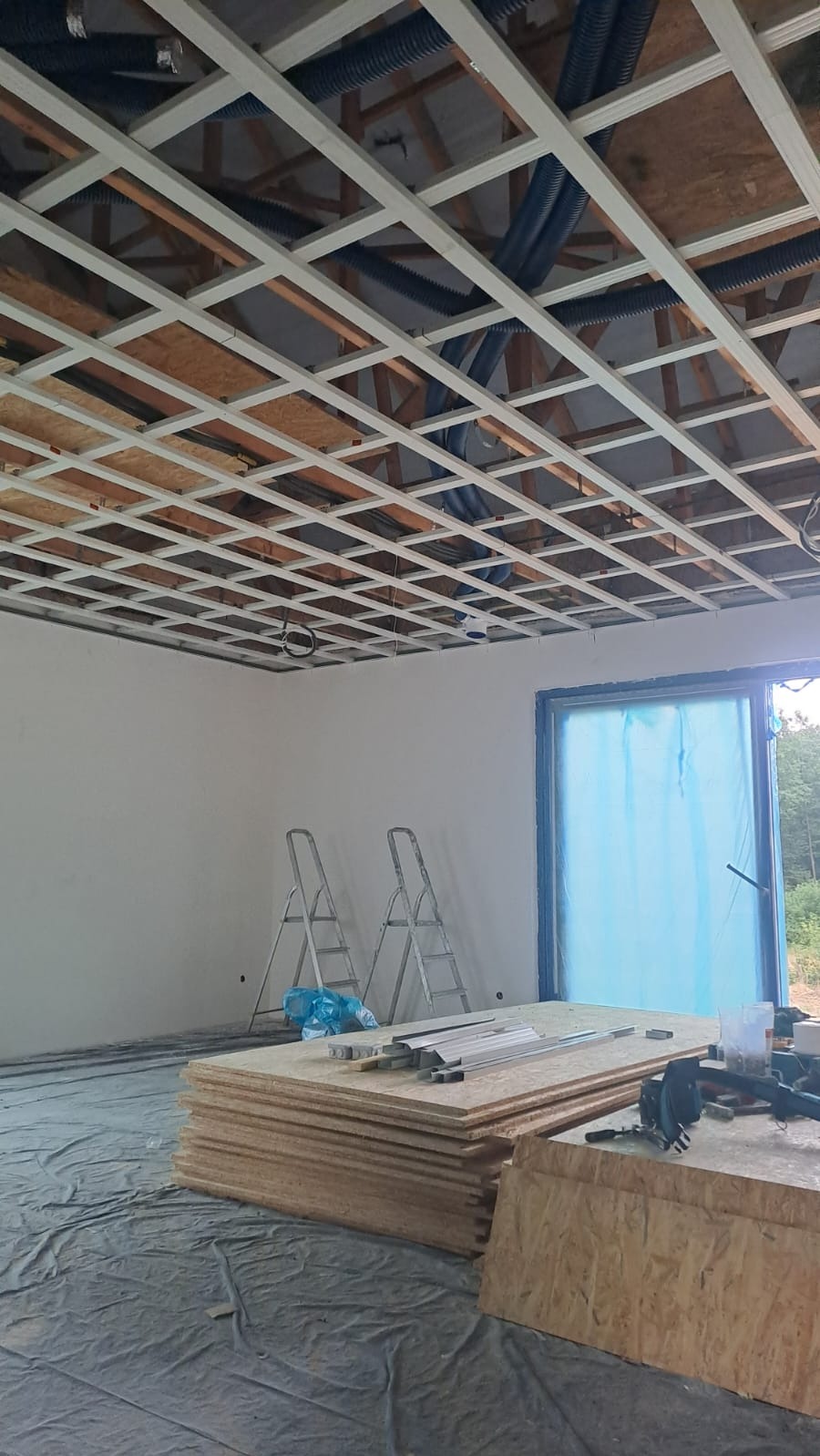 Montaż sufitu podwieszanego z widoczną konstrukcją metalową i izolacją. Dwie drabiny opierają się o ścianę. Na podłodze leżą płyty OSB i narzędzia.