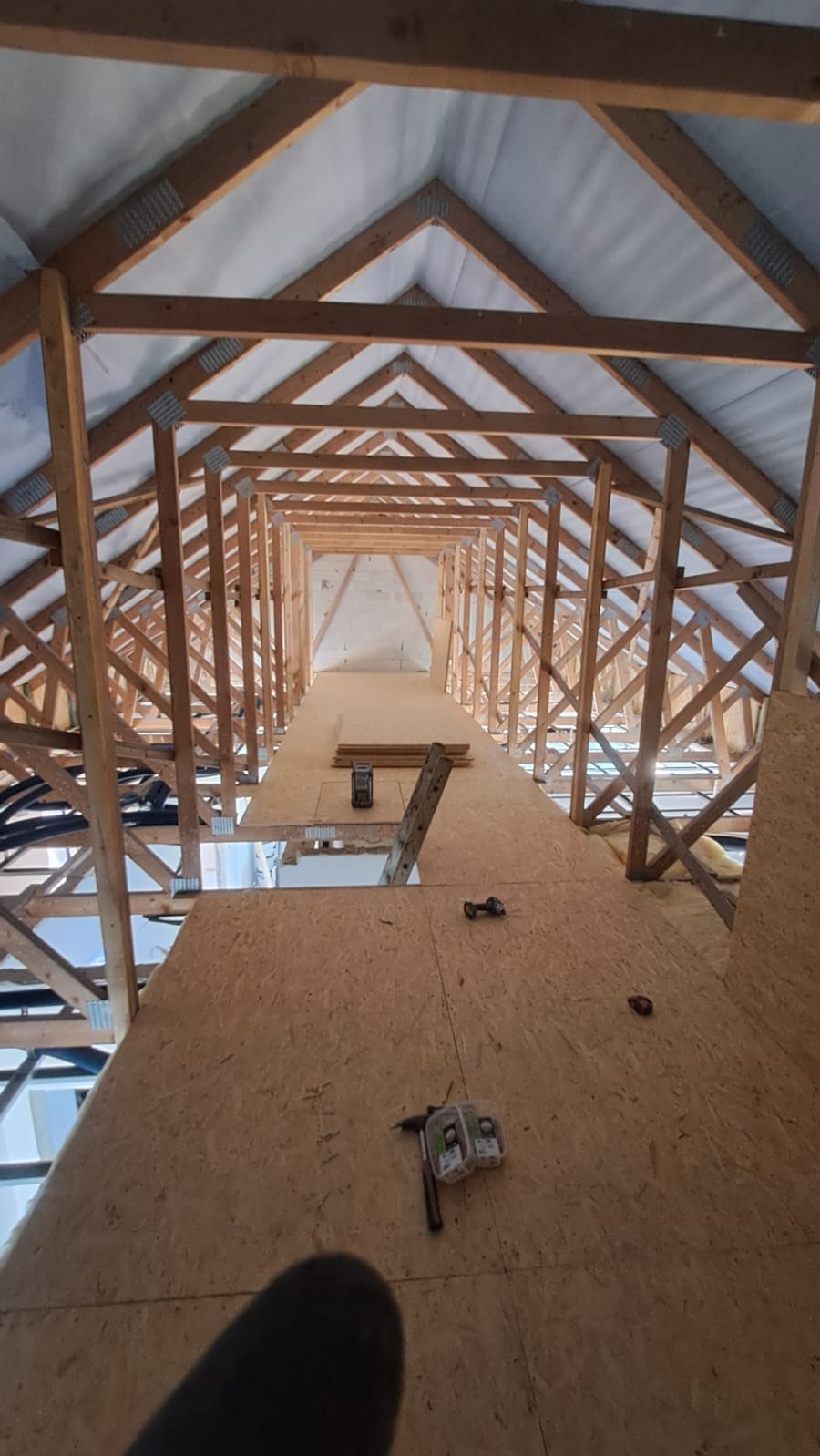 Szeroki kadr na drewnianą konstrukcję dachu z płytami OSB, widok z dołu. Na płytach leżą narzędzia i akcesoria montażowe. W tle widoczna drabina i oświetlenie.