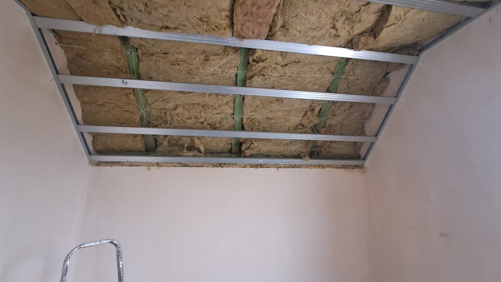 Sufit podwieszany w trakcie montażu: metalowa konstrukcja z izolacją z wełny mineralnej, widoczne elementy mocujące. Ściany w jasnym kolorze, drabina w rogu.