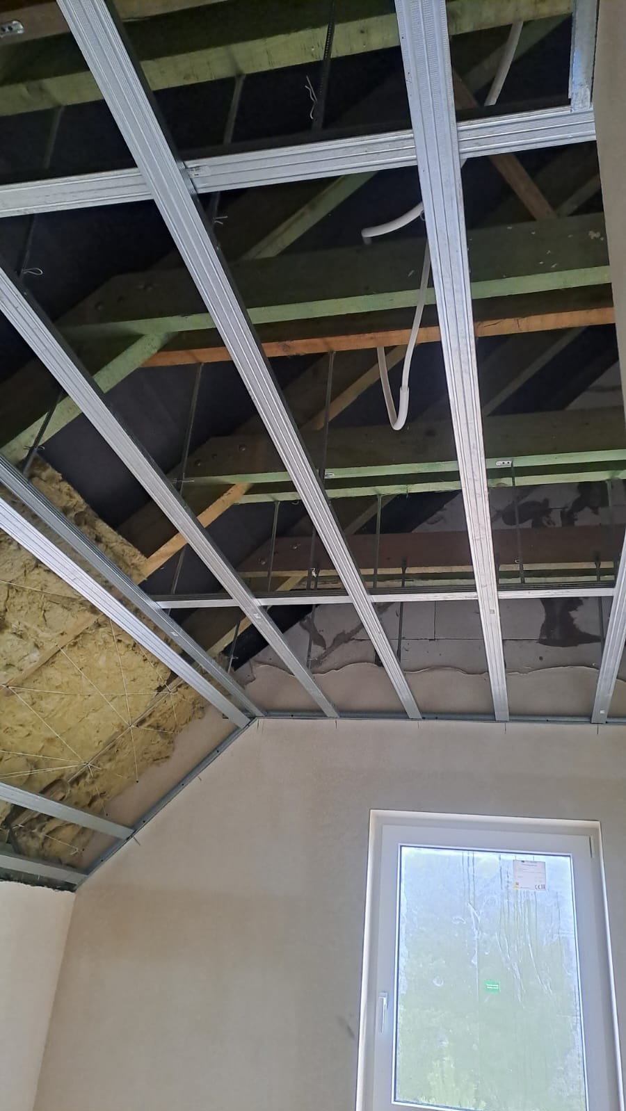 Montaż metalowego stelażu sufitu podwieszanego z widoczną izolacją i fragmentem okna. Prace wykończeniowe w pomieszczeniu o skośnym suficie.