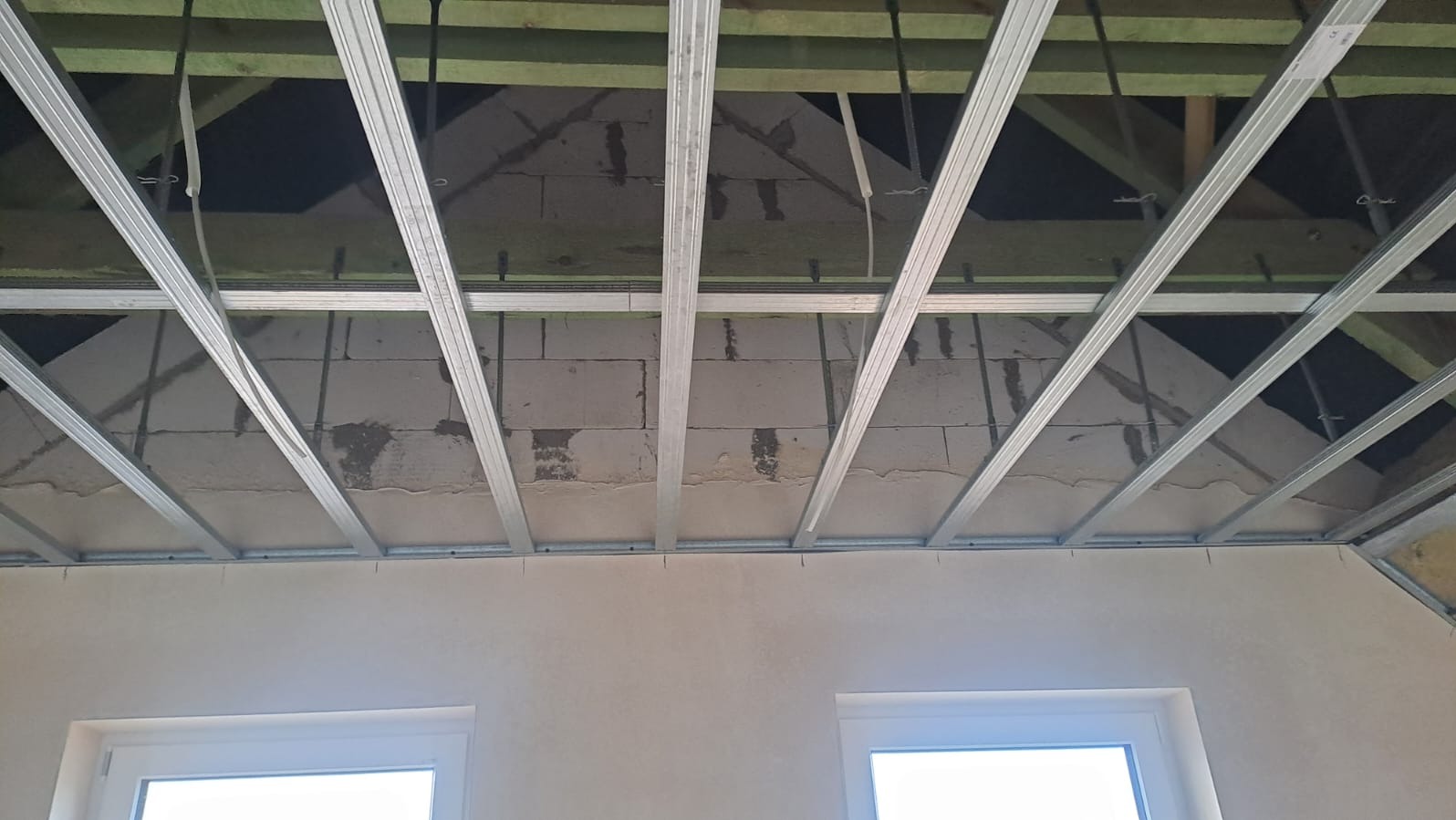 Metalowa konstrukcja sufitu podwieszanego z widocznymi elementami drewnianymi więźby dachowej, przygotowana do montażu płyt. Widoczne okna i fragment ściany.