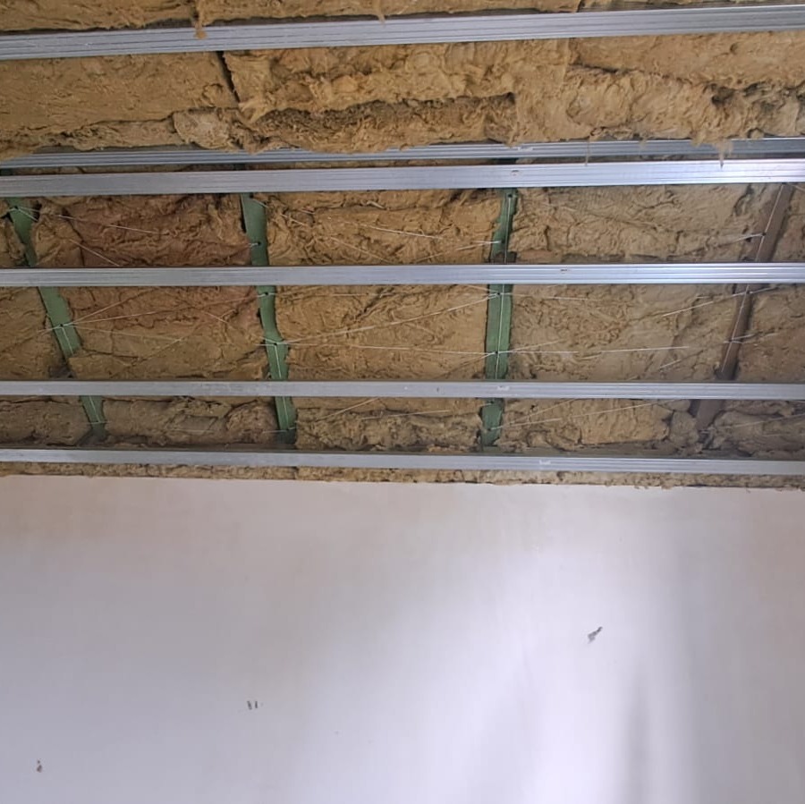 Montaż izolacji z wełny mineralnej na metalowym ruszcie sufitowym, widoczne zielone elementy mocujące. Ściana w tle przygotowana do dalszych prac wykończeniowych.