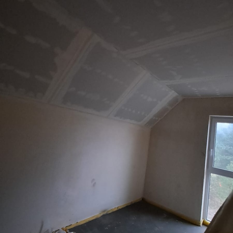 Poddasze w trakcie prac wykończeniowych: zamontowane płyty gipsowo-kartonowe na skośnym suficie, widoczne łączenia i szpachlowanie. Okno w tle.