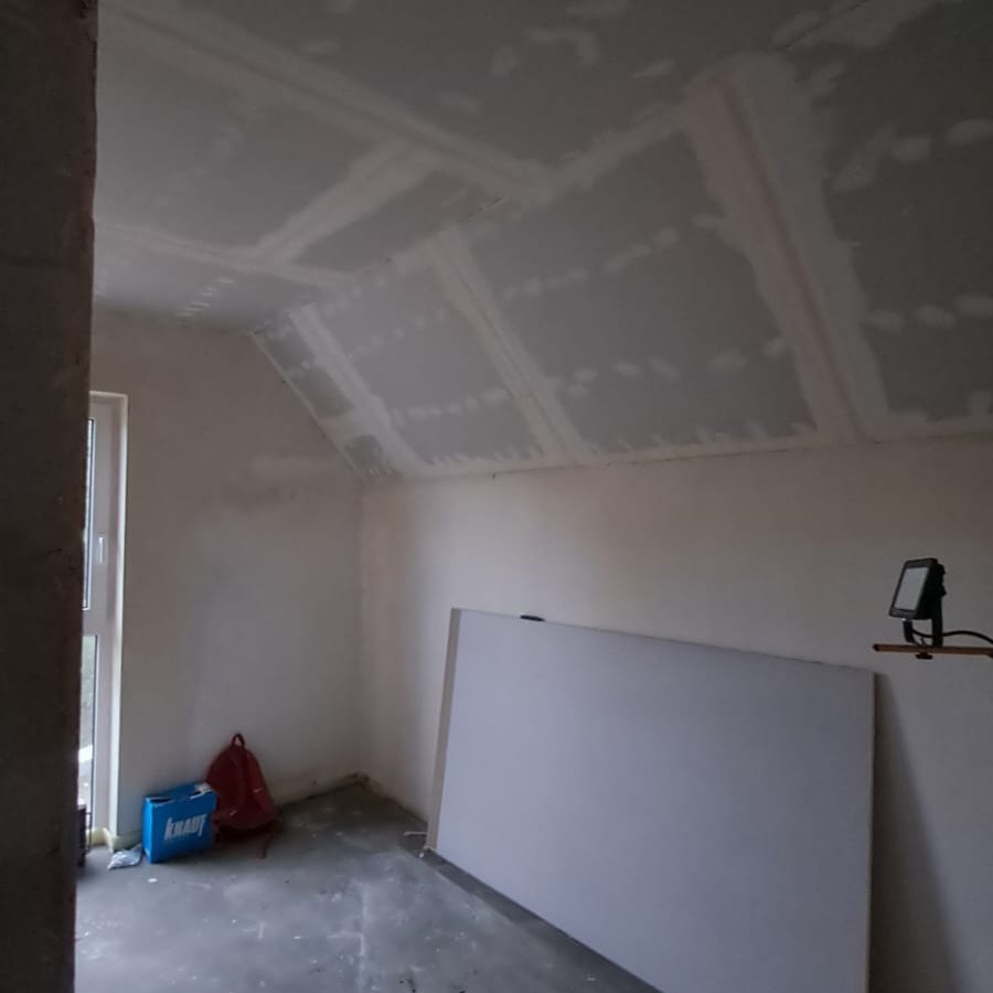 Poddasze po montażu płyt gipsowo-kartonowych na skośnym suficie, widoczne łączenia i szpachlowanie. Na podłodze leży płyta i stoi lampa budowlana.