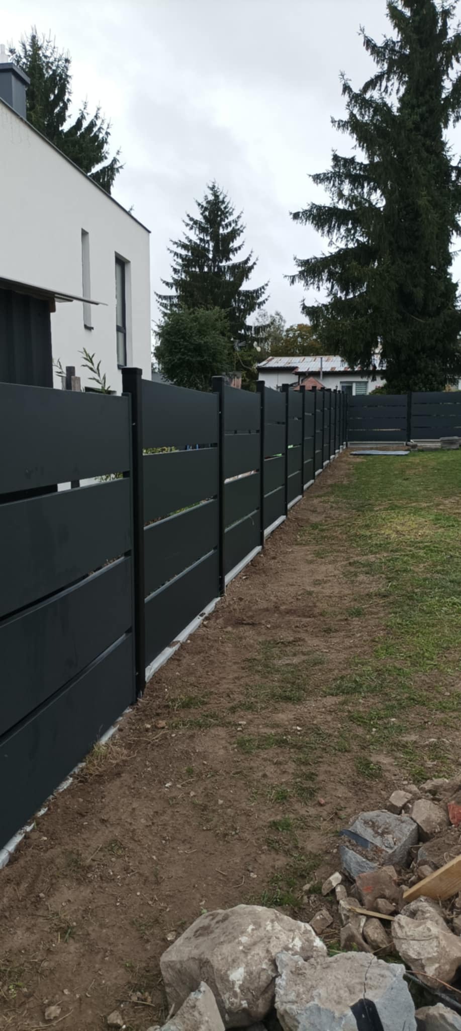 Nowoczesne, poziome ogrodzenie panelowe w kolorze antracytowym, oddzielające posesję od sąsiadów. Widok z perspektywy trawnika, z fragmentem domu w tle.