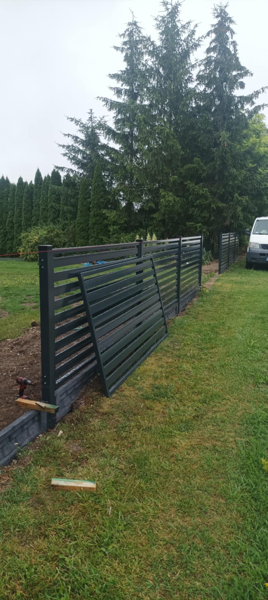 Montaż nowoczesnego ogrodzenia panelowego w kolorze grafitowym. Widoczne słupki, panele i narzędzia. Tło: zielony trawnik i wysokie drzewa.