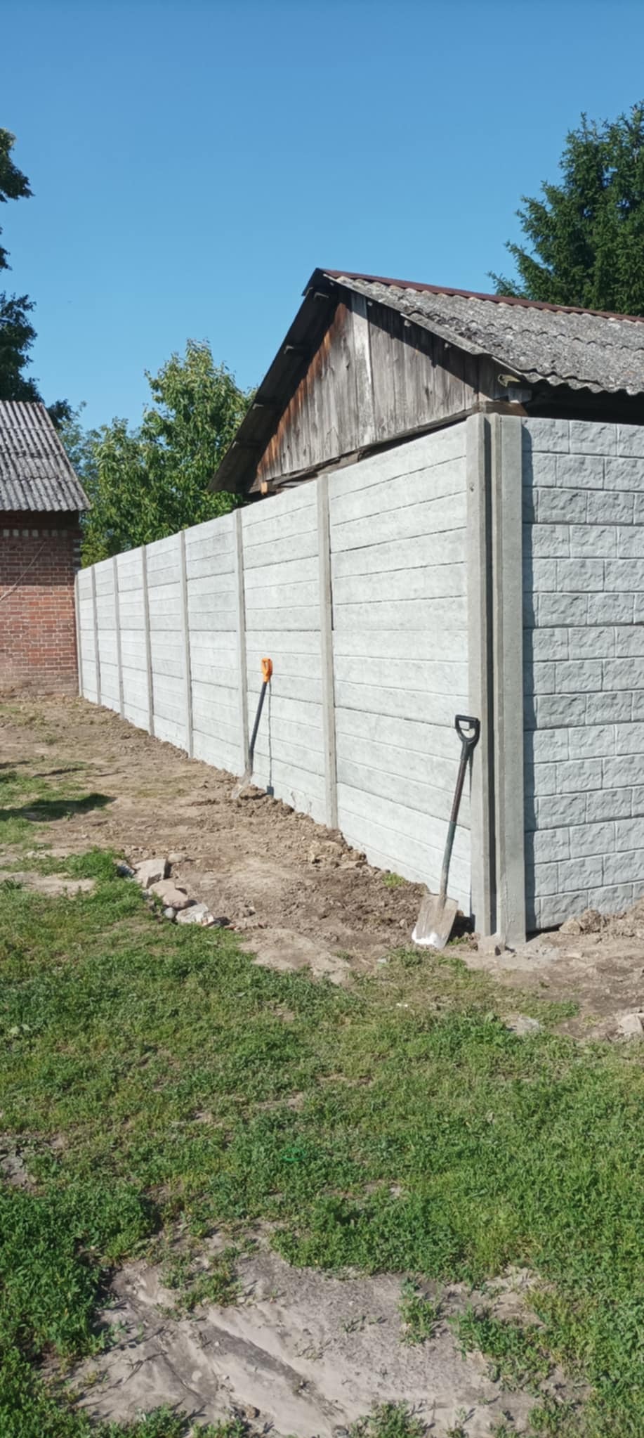 Długi, szary, betonowy płot z fakturą cegły, oddzielający posesję. W tle budynki i drzewa. Dwie łopaty oparte o płot. Słoneczny dzień.