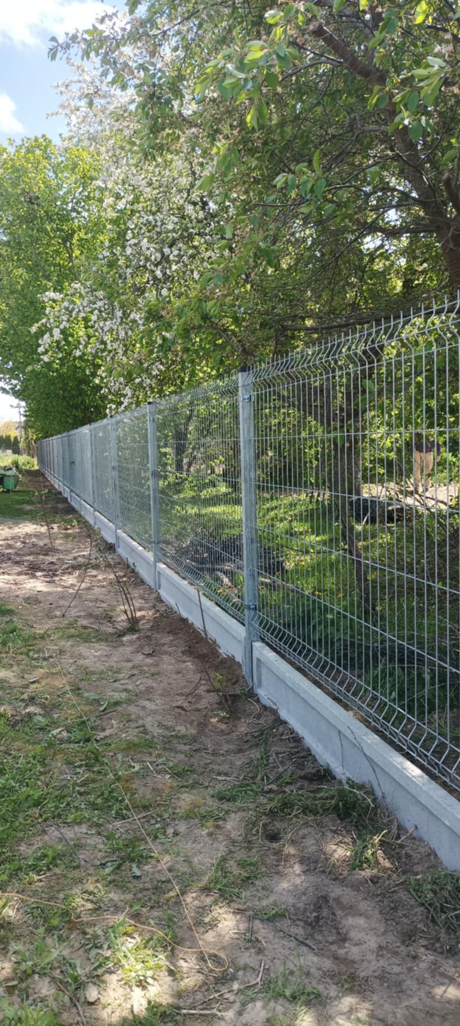 Długi, szary panel ogrodzeniowy na betonowej podmurówce w Strzyżewicach, z zielenią w tle i kwitnącymi drzewami. Widok z boku, w słoneczny dzień.