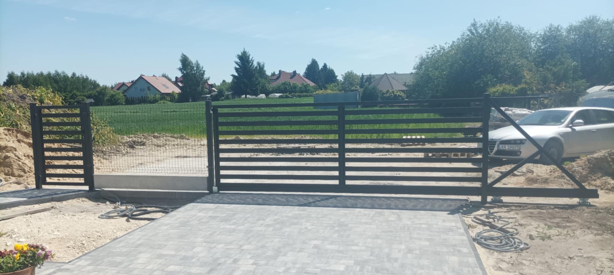Nowoczesna, przesuwna brama metalowa w kolorze antracytu, z poziomymi profilami, zamontowana na posesji z kostką brukową. W tle widoczny dom i trawnik.