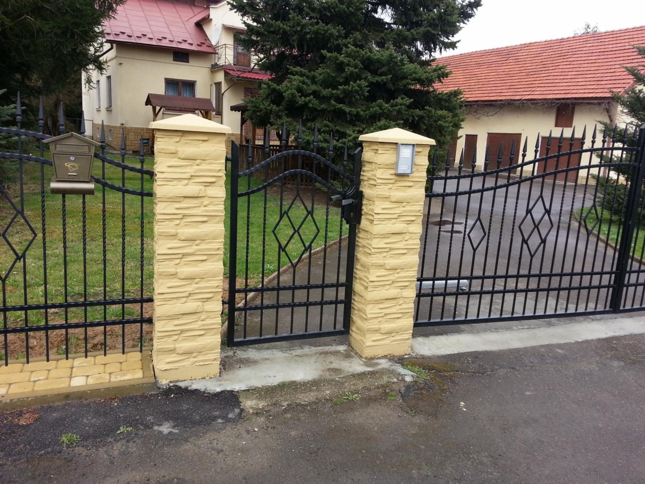 Czarna, metalowa brama wjazdowa i furtka z dekoracyjnymi rombami, osadzona na żółtych, teksturowanych słupkach imitujących kamień, z widoczną skrzynką na listy i domofonem.