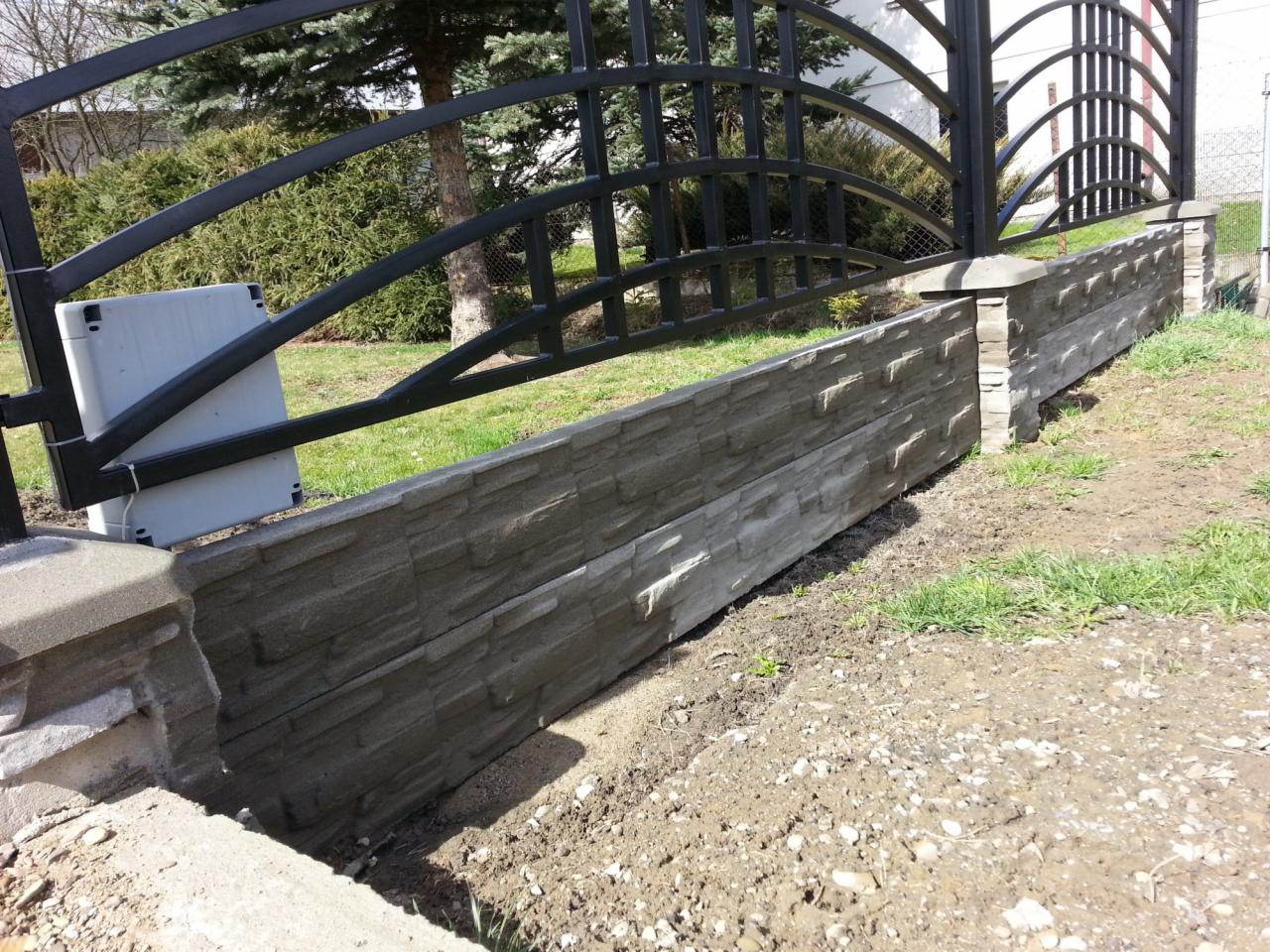 Fragment ogrodzenia z betonowych elementów imitujących kamień, połączony z metalową bramą wjazdową z automatyką, widoczne trawnik i drzewa w tle.