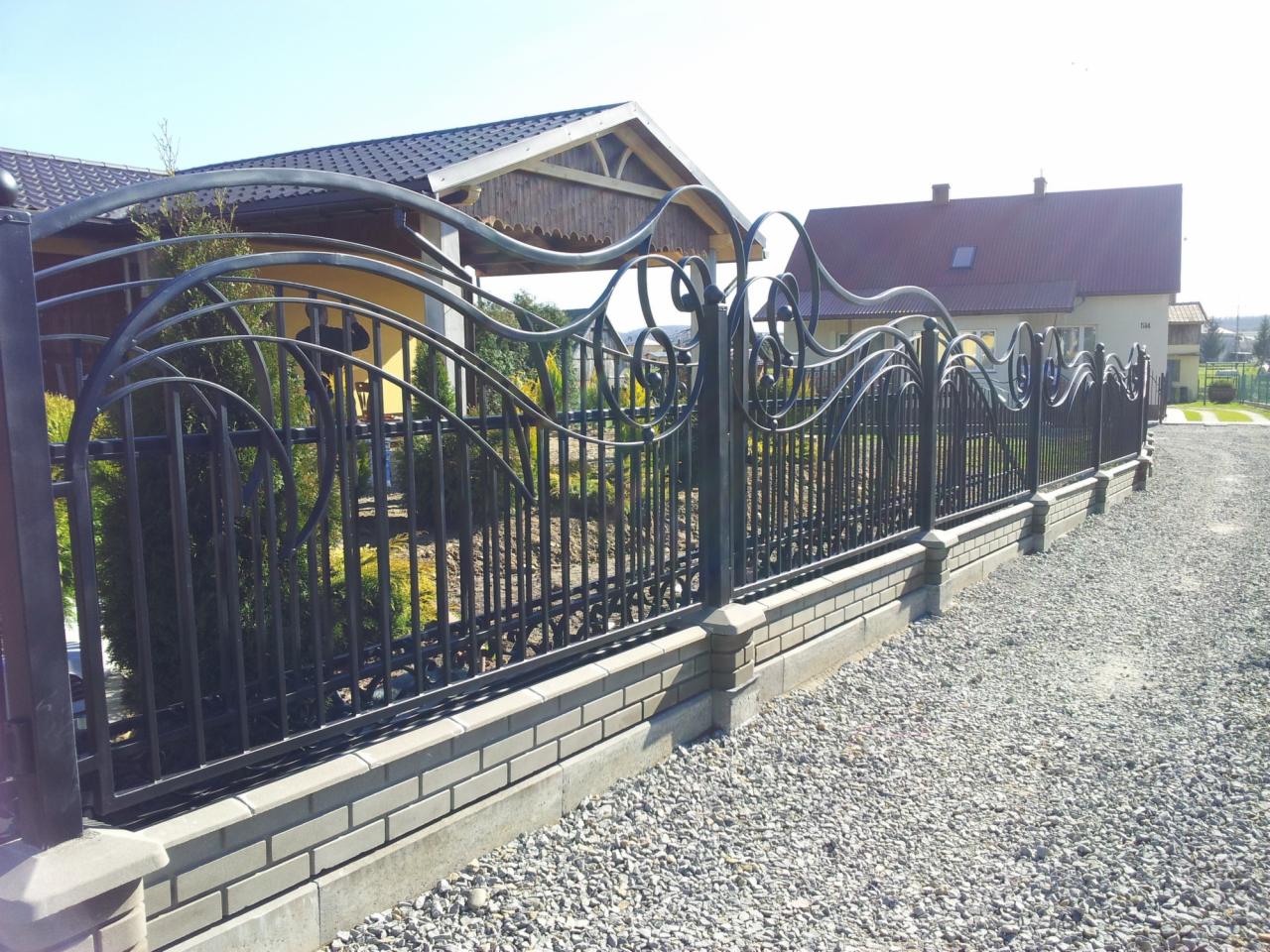 Eleganckie, czarne ogrodzenie metalowe z ozdobnymi elementami, osadzone na niskim murku z szarej cegły, na tle domu z żółtą elewacją i żwirowego podjazdu.