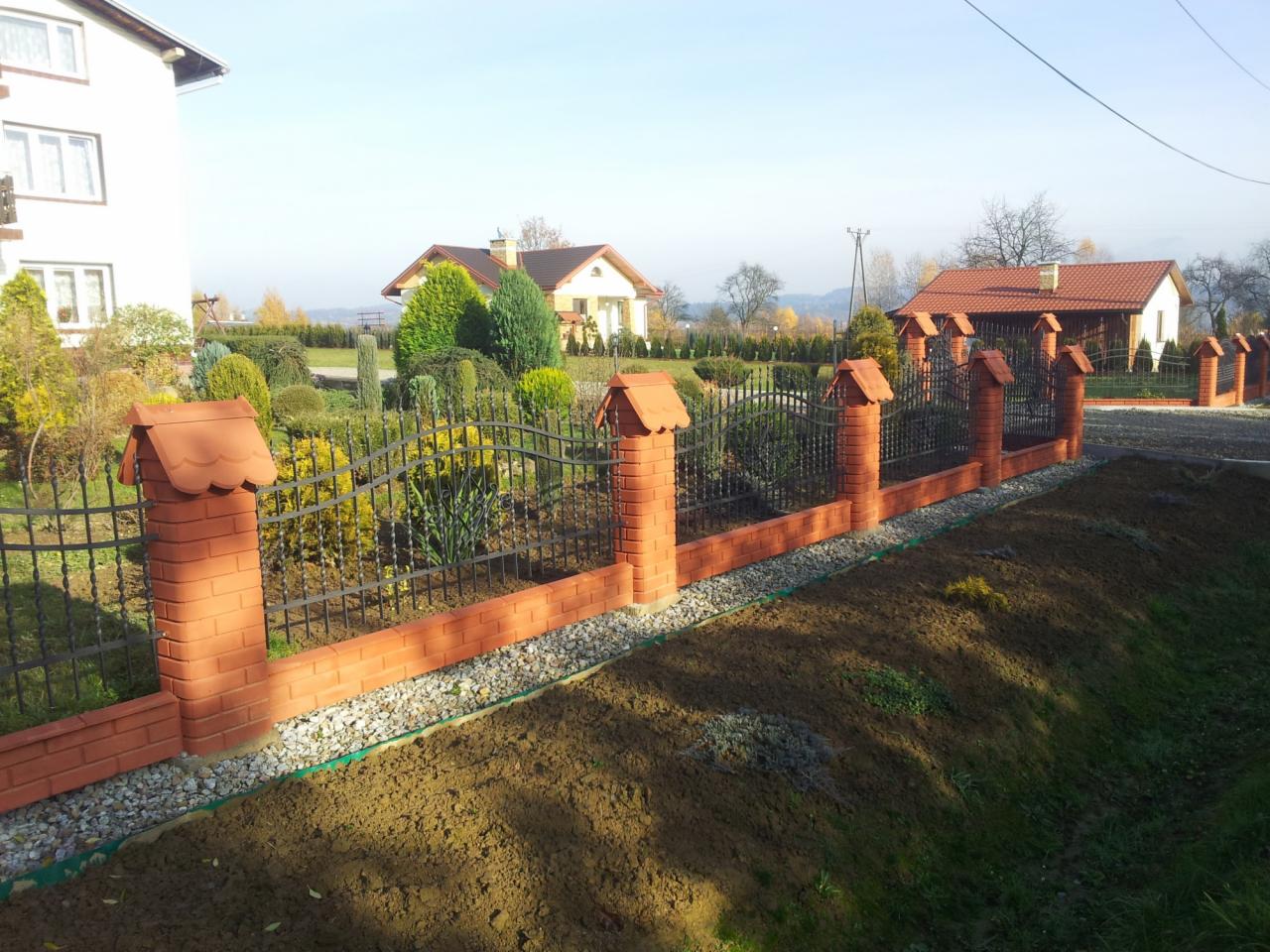 Nowoczesne ogrodzenie metalowe z elementami murowanymi z cegły klinkierowej, zwieńczone daszkami, oddzielające posesję z zadbanym ogrodem od niezagospodarowanego terenu.