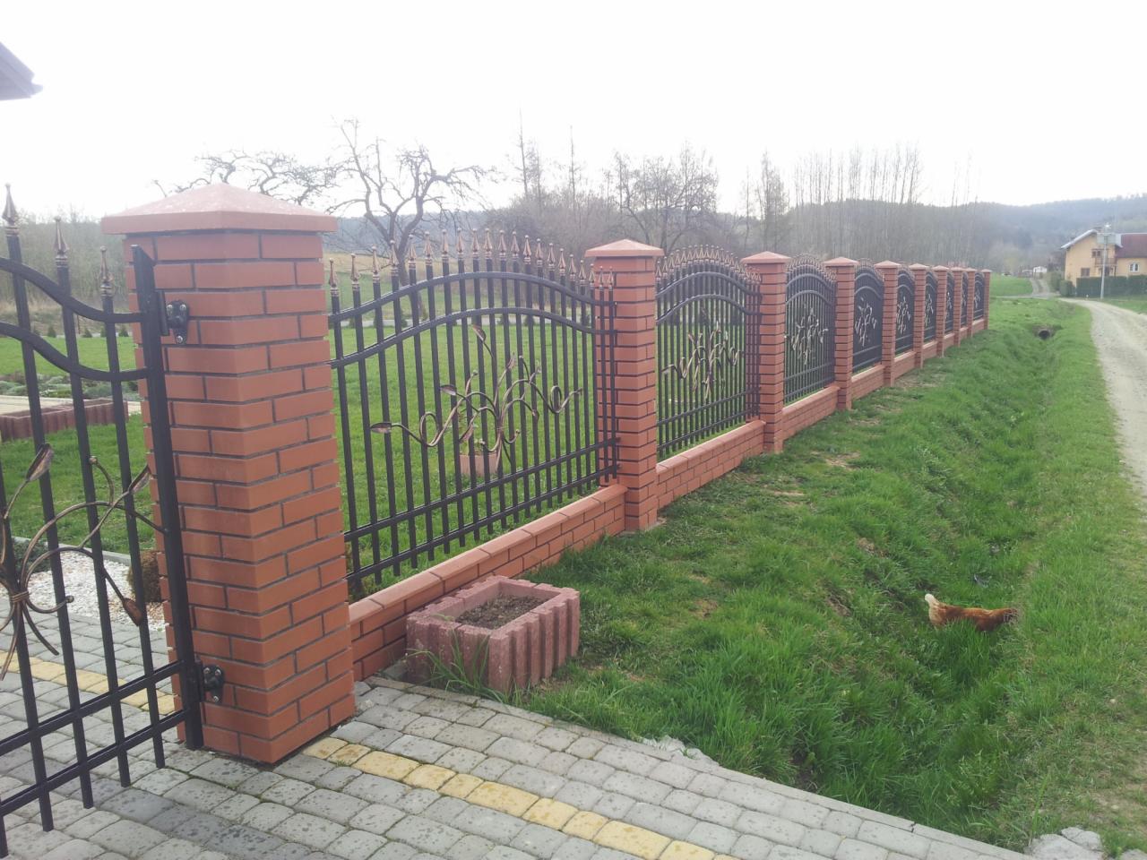 Długie ogrodzenie z metalowymi przęsłami i ceglanymi słupkami, widok z boku, na trawniku spaceruje kura.