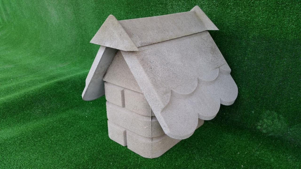 Mała, betonowa budka dla ptaków z dekoracyjnymi, zaokrąglonymi krawędziami dachu, umieszczona na sztucznej, zielonej trawie.