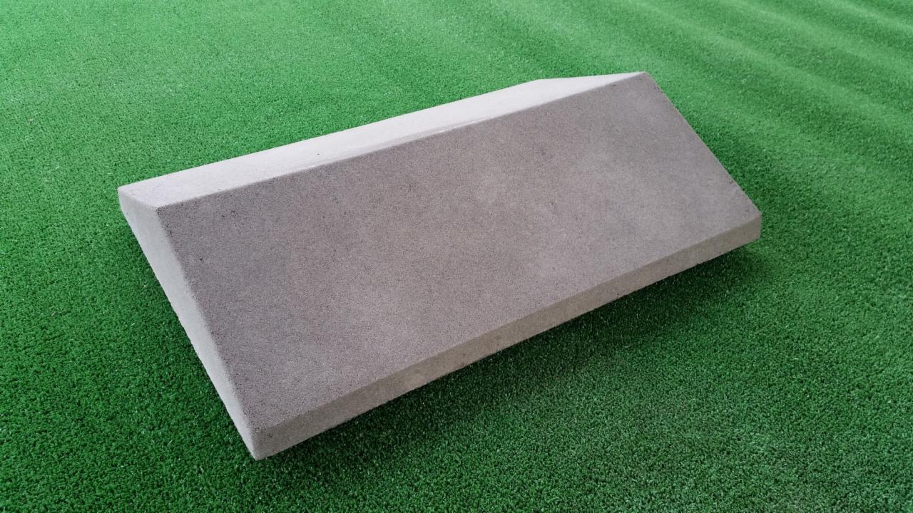 Pojedynczy, szary, betonowy prefabrykat na tle sztucznej, zielonej trawy, o trapezoidalnym przekroju.